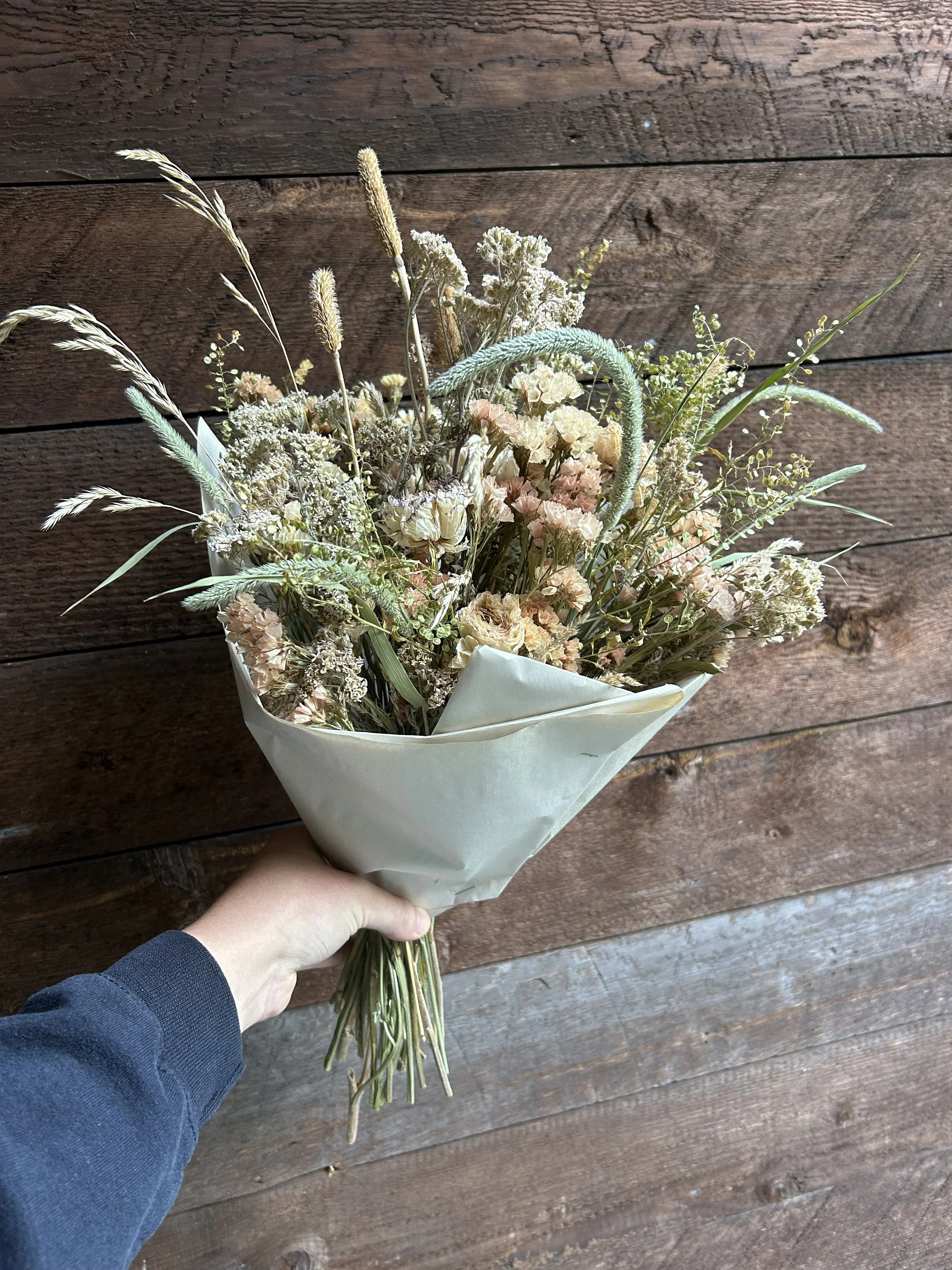 Hand holding a bouquet of dried flowers wrapped in paper, with a wooden wall background.