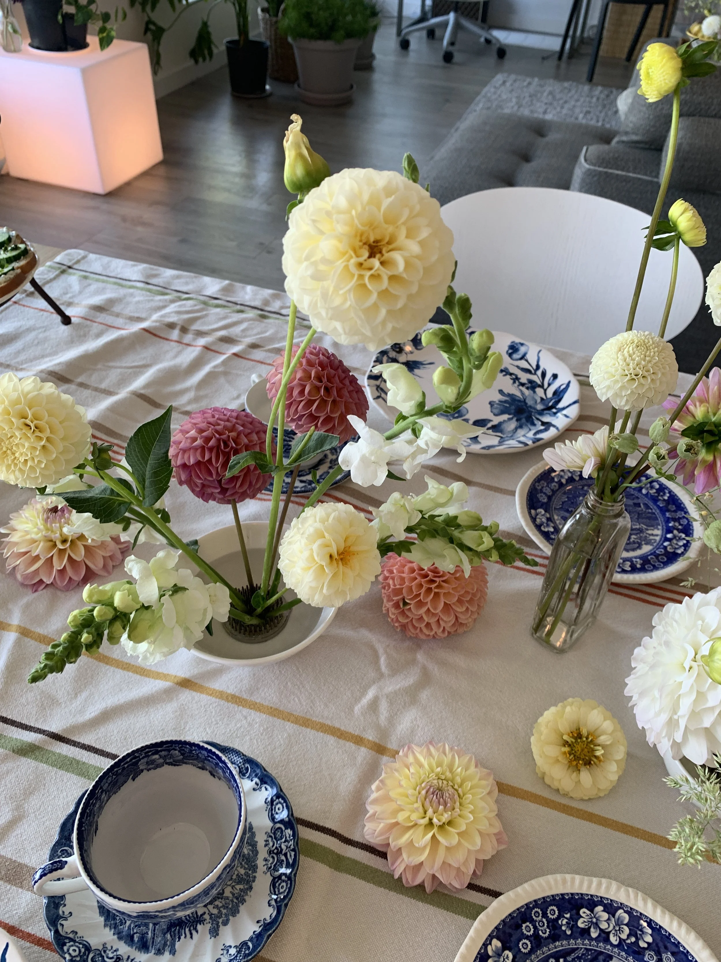 A table decorated with a variety of colorful flowers in vases and bowls, including white, pink, and yellow blooms, along with blue and white patterned dishware, set in a bright room with hardwood floors and potted plants.