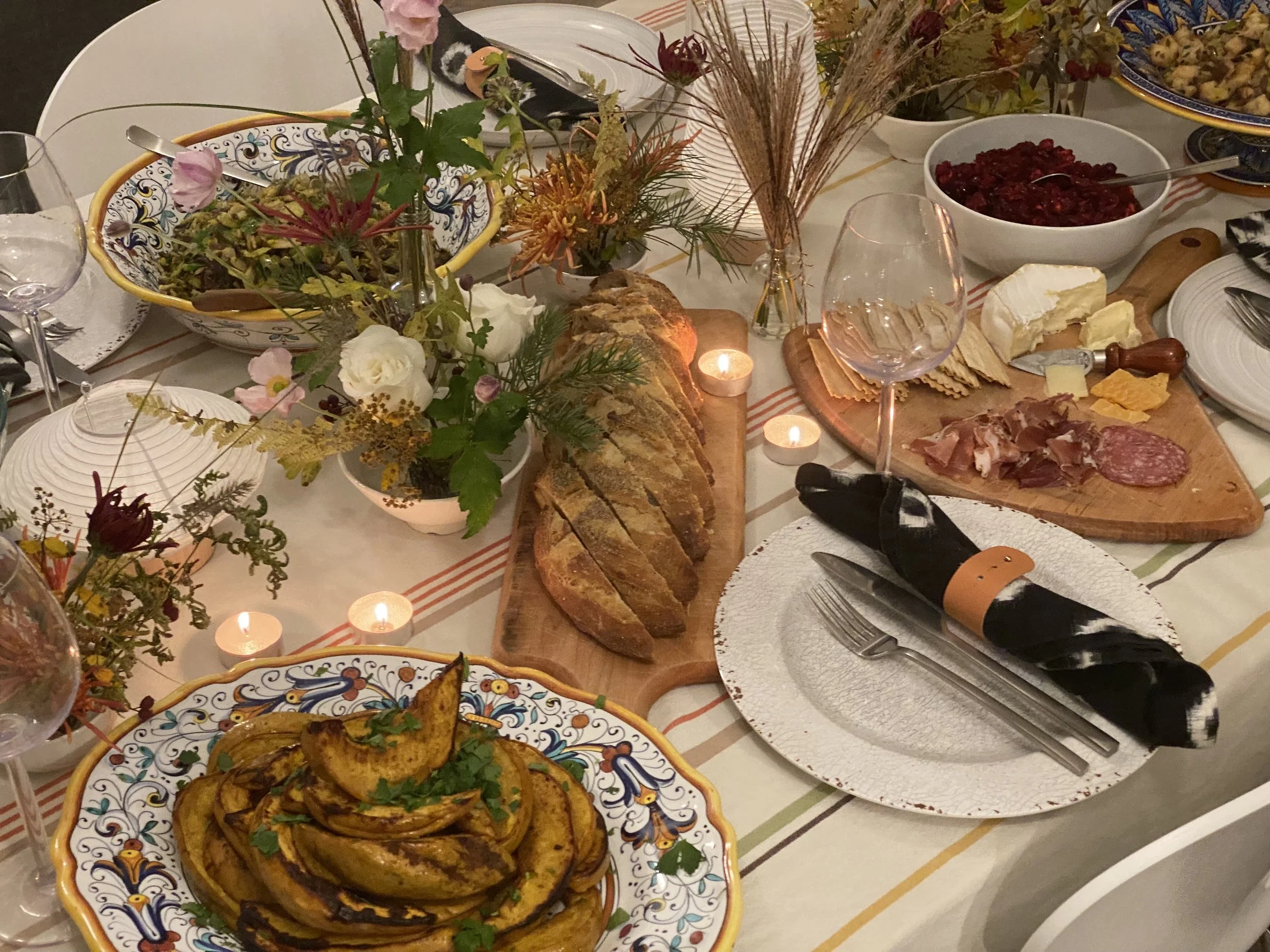 A festive dinner table set with various dishes including roasted peppers, a bread loaf, cheeses, sliced cured meats, a bowl of cranberries, and a salad. The table is decorated with flowers and small candles.