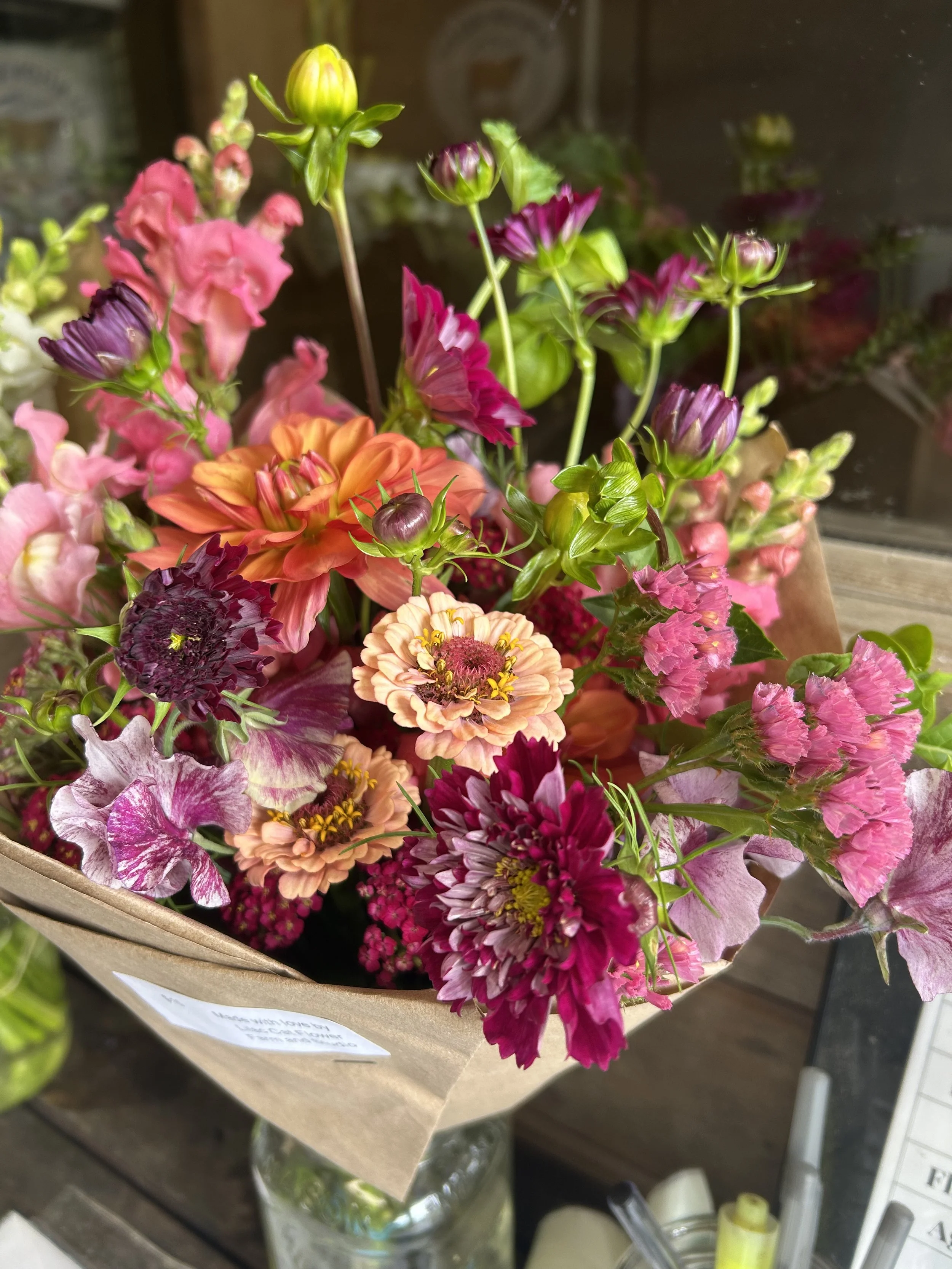 A colorful bouquet of various flowers including pink, purple, orange, and peach, wrapped in brown paper and placed in a glass jar.