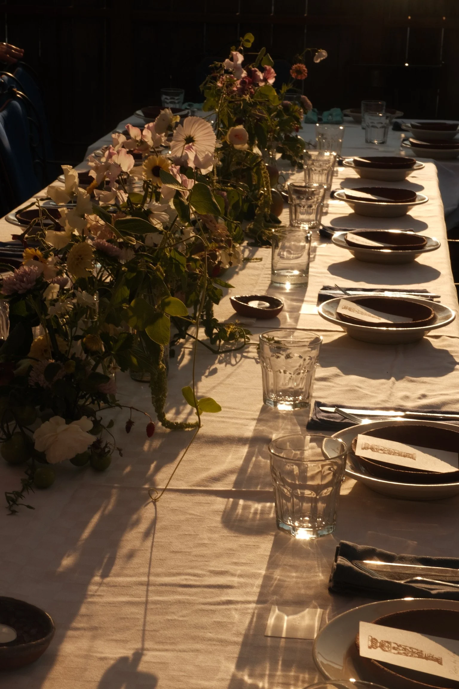 Long dining table with floral centerpiece, glassware, plates, and napkins arranged for a meal, illuminated by warm sunlight.