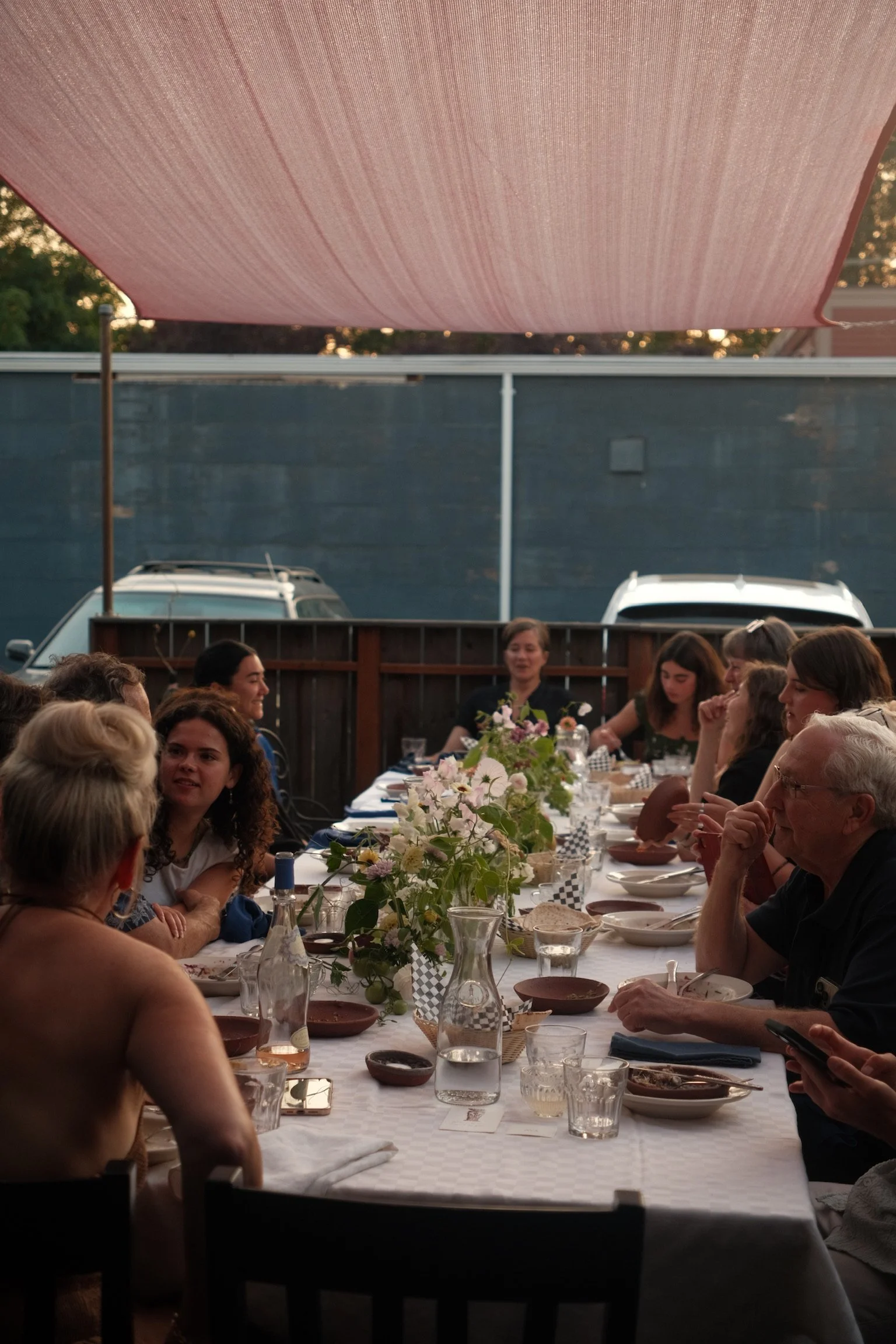 People gathered around a long outdoor dining table under a pink shade sail, with plates, glasses, and floral centerpieces, during a social gathering in the evening.
