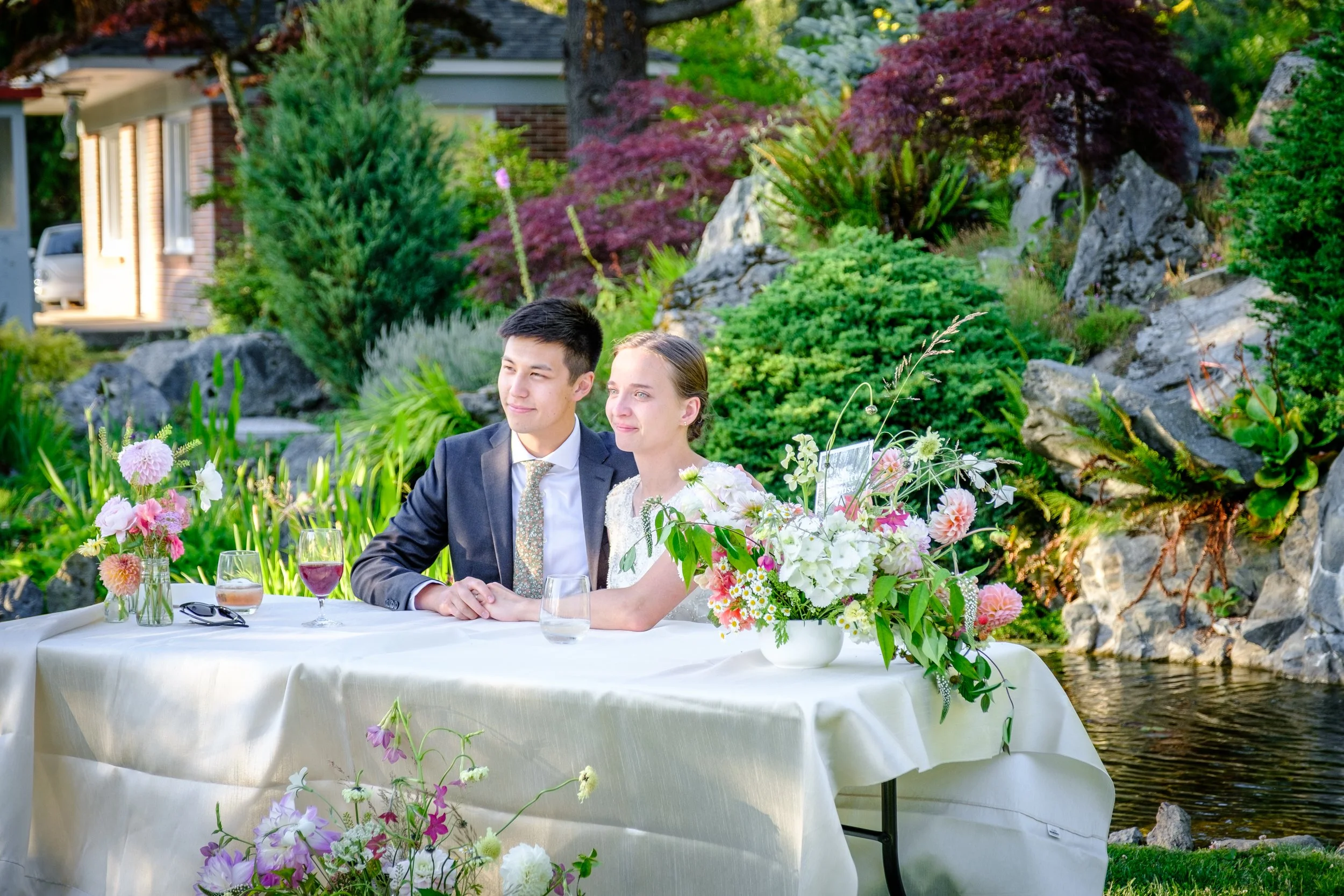 A young couple at a wedding reception seated at a table outdoors with a lush garden and pond in the background. The table is decorated with floral arrangements in vases and glasses of drinks.