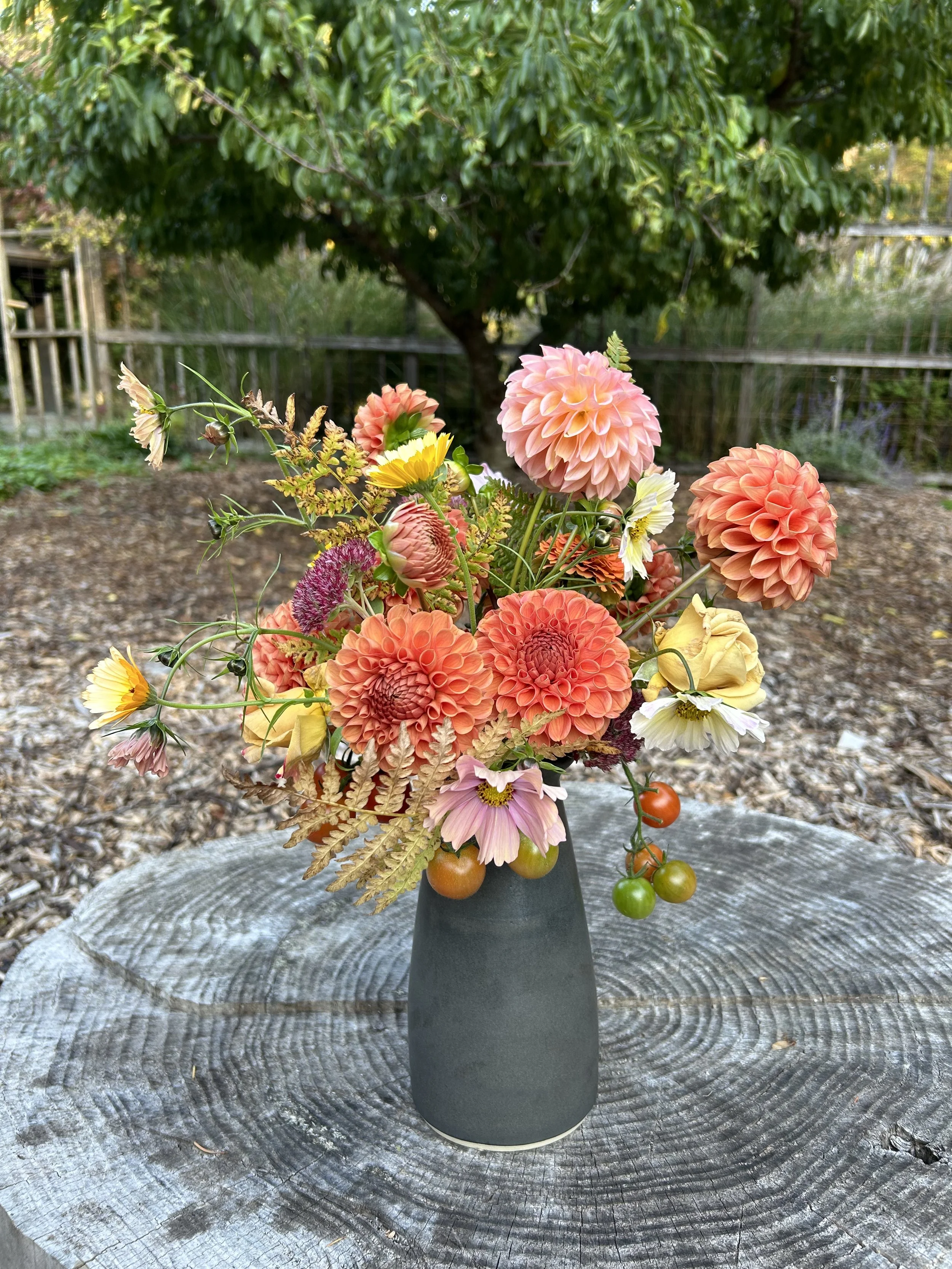A bouquet of multicolored flowers including dahlias, cosmos, and other garden flowers in a dark gray vase on a wooden surface outdoors with a tree and wooden fence in the background.