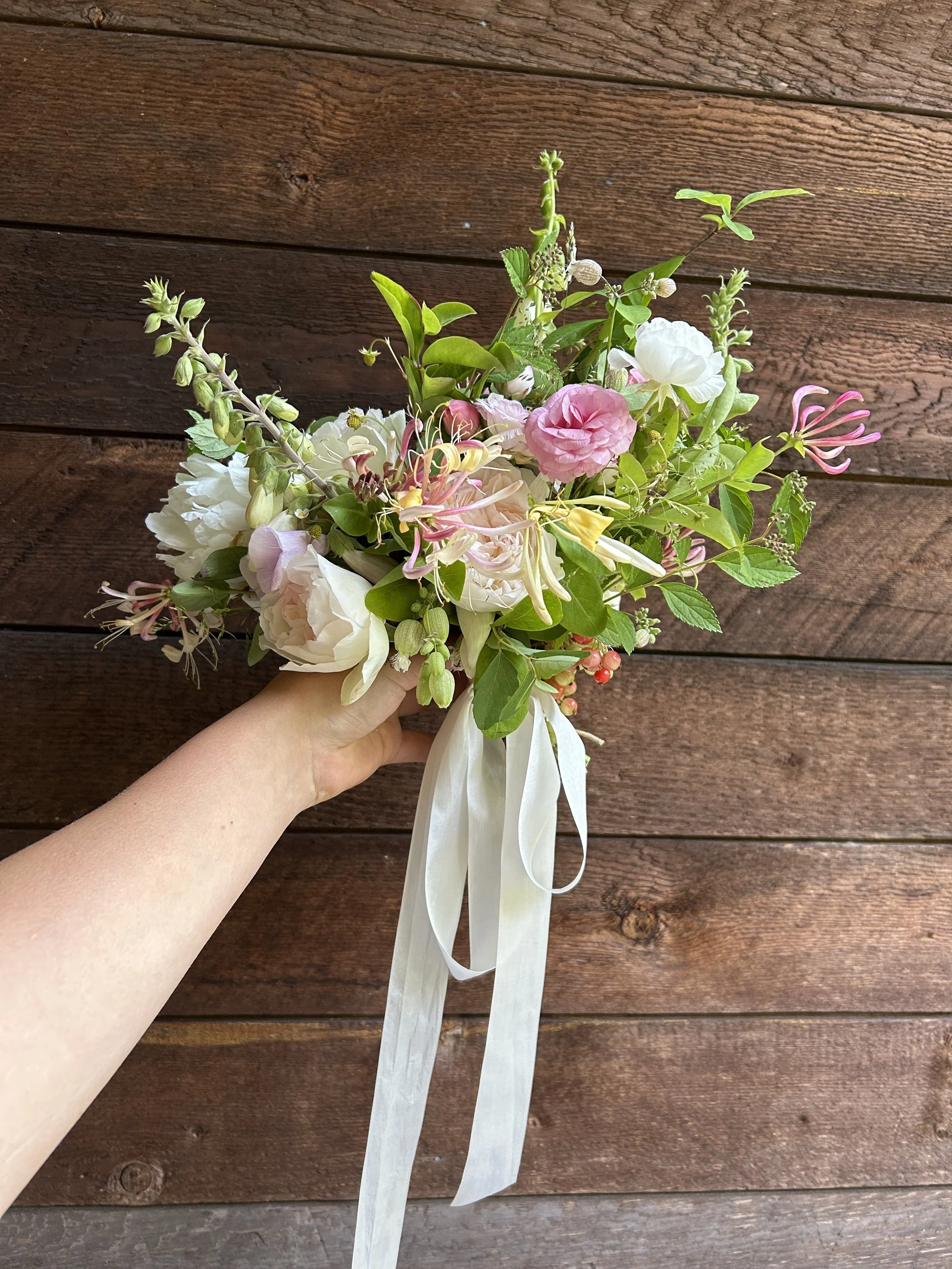 A hand holding a bouquet of mixed pink, white, and green flowers and leaves against a wooden background.