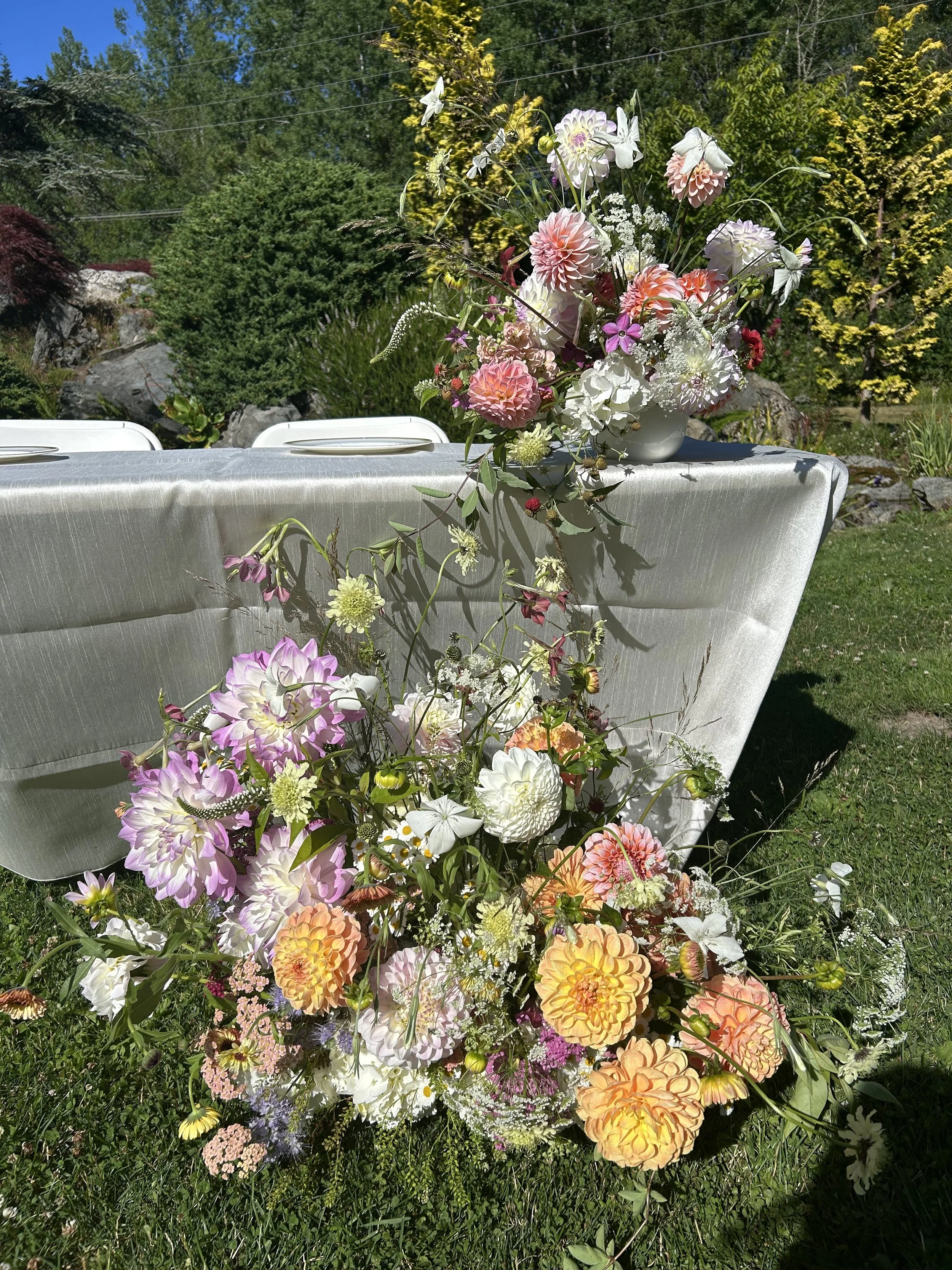 Colorful flower arrangements on a table outdoors with greenery and trees in the background.