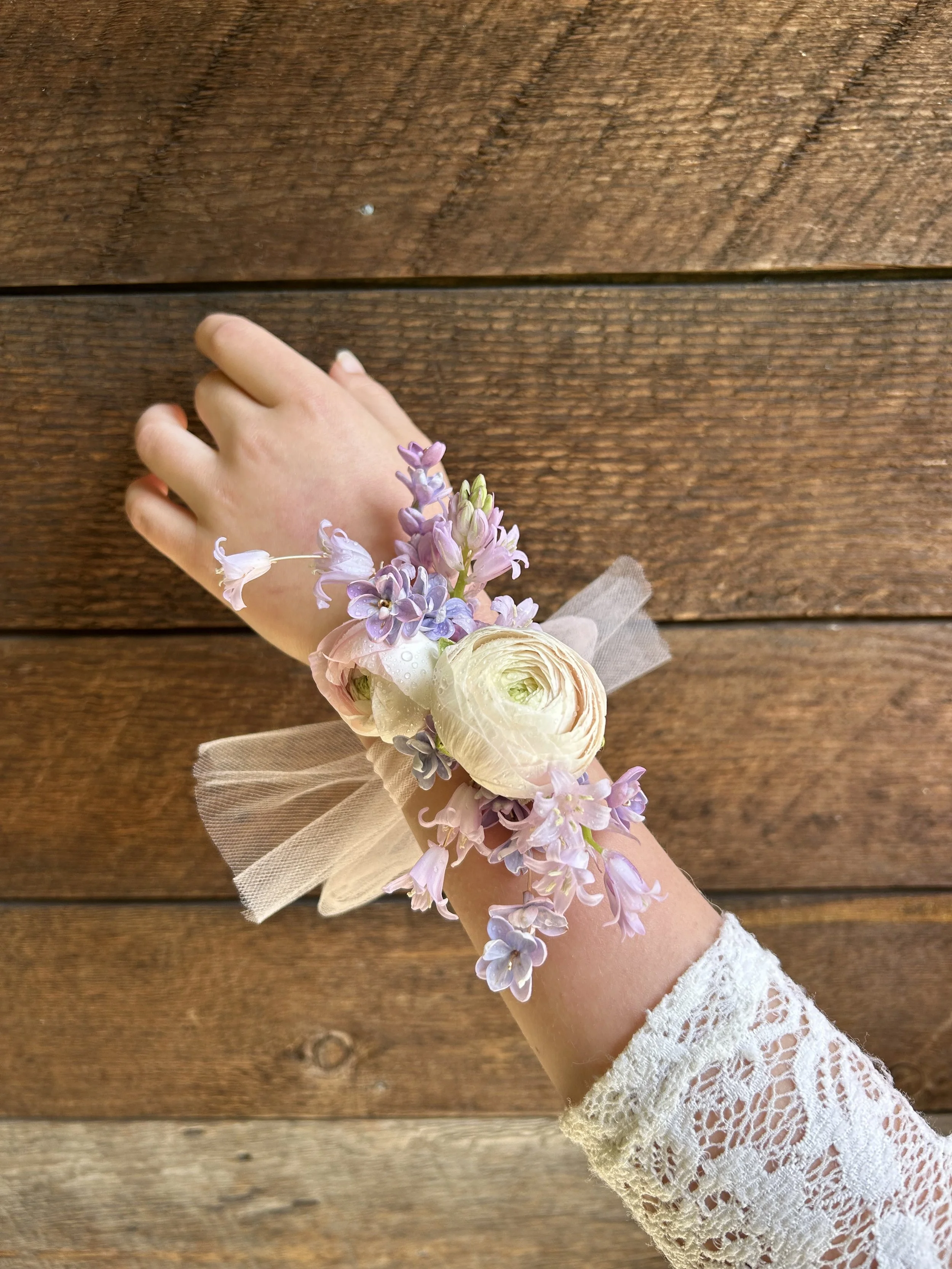 A hand wearing a lace sleeve with a floral wrist corsage consisting of purple, pink, and white flowers, and beige ribbon, against a wooden background.