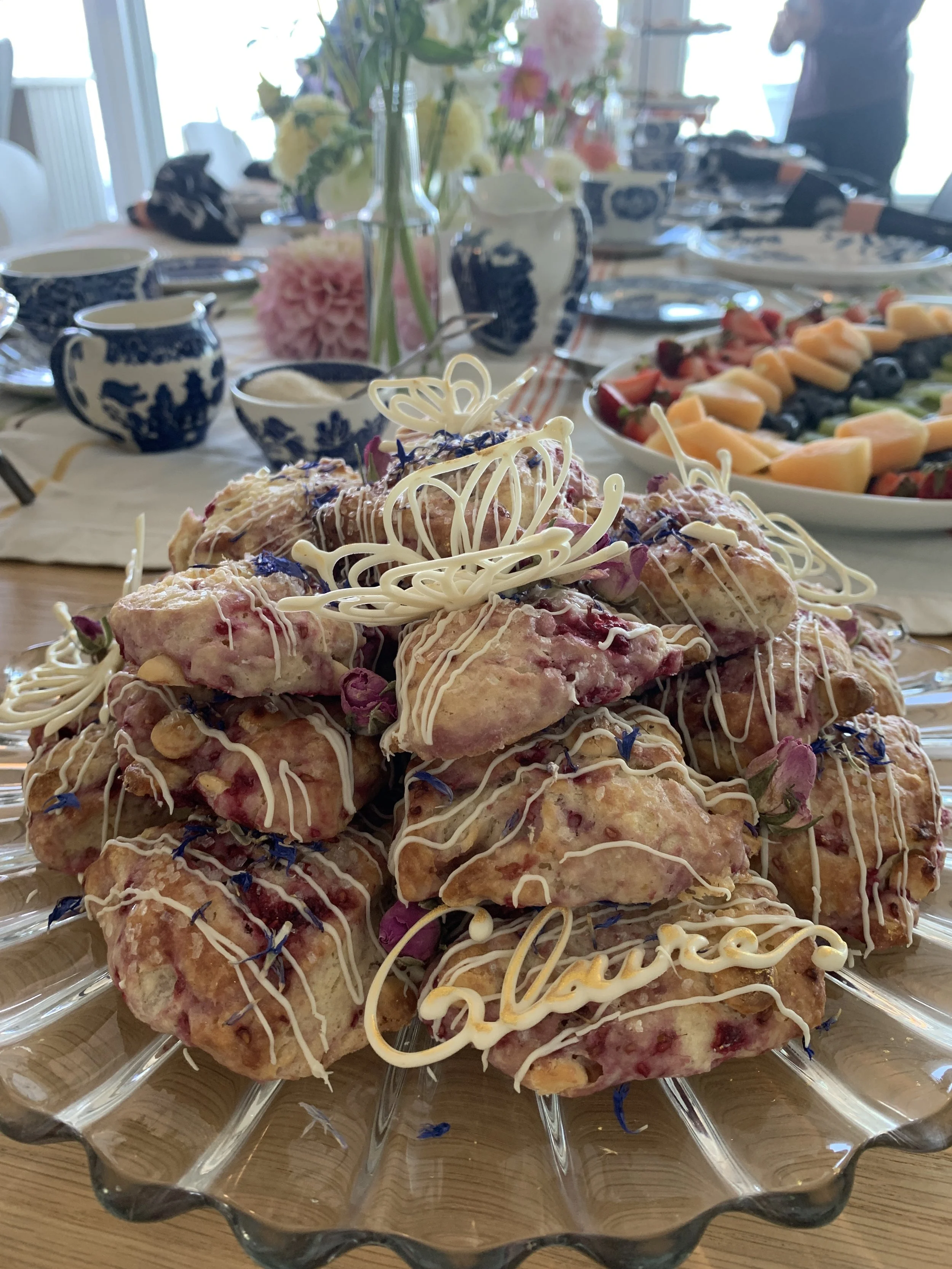 Plate of decorated cookies with white chocolate writing and flowers, set on a dining table with fruit, floral arrangements, and blue and white dishware in the background.