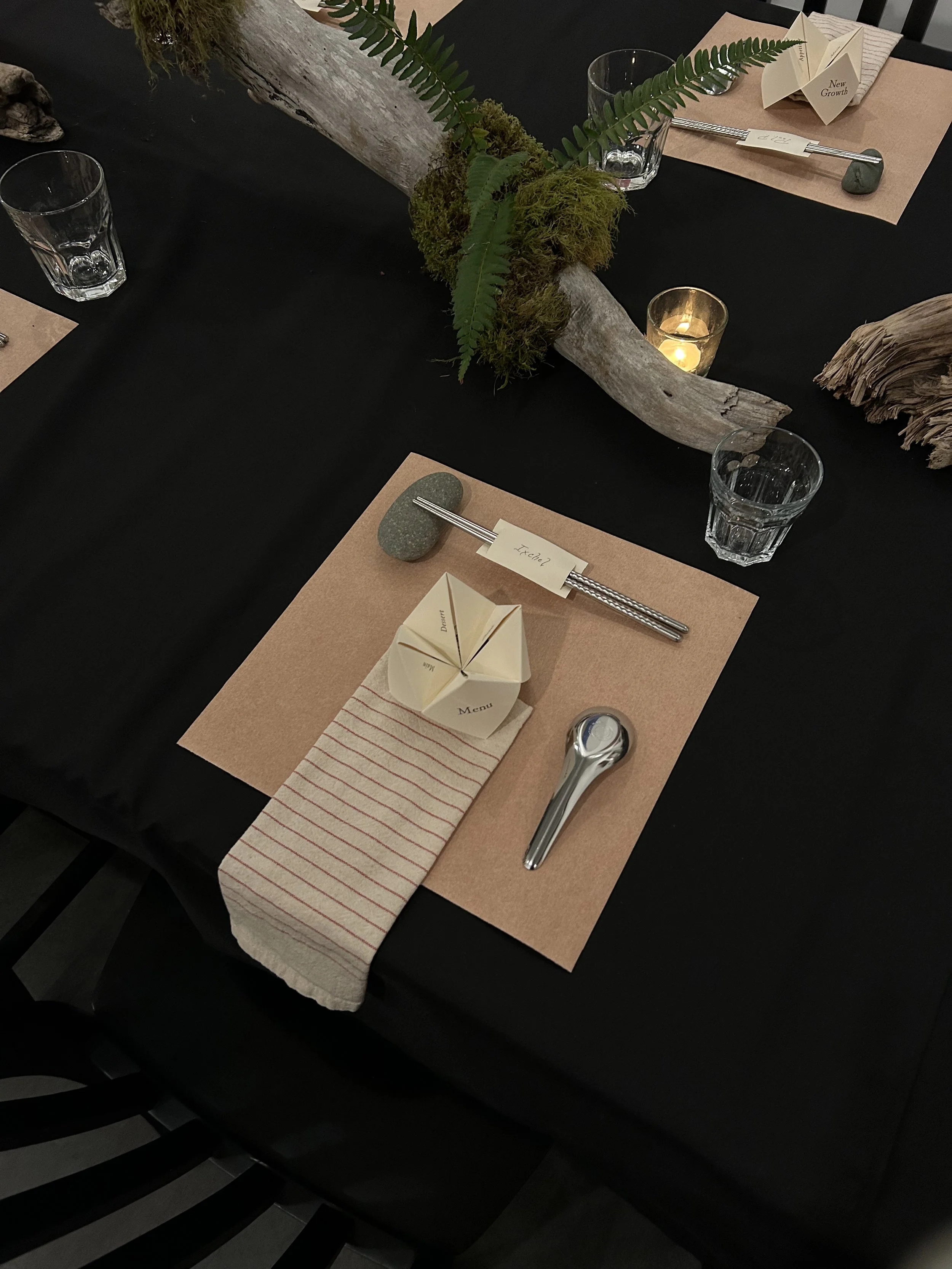 Table setting with brown placemats, a folded napkin, a menu card, and utensils, decorated with a driftwood piece, moss, and fern leaves, with glassware and a candle.