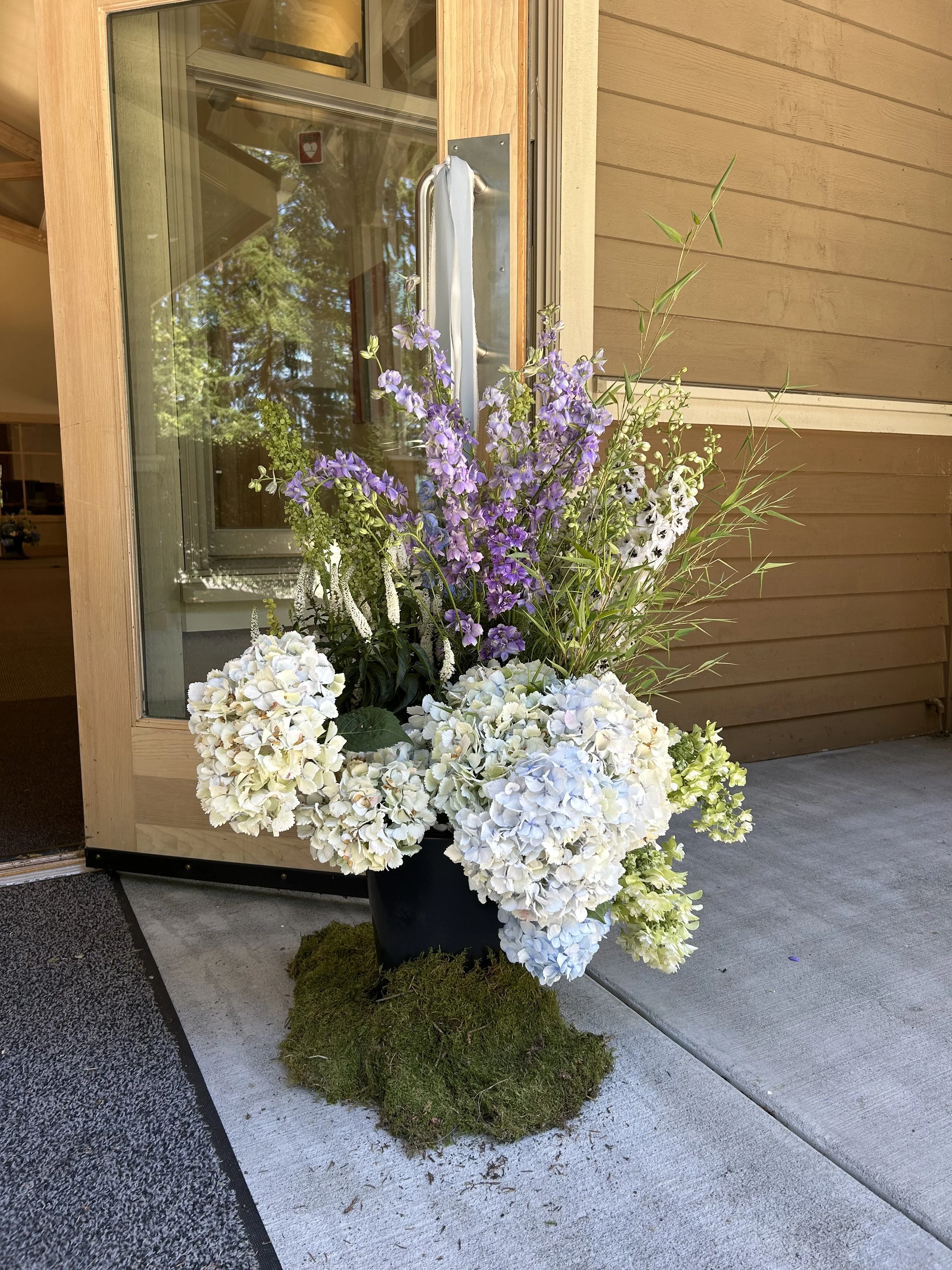 A large black flower pot filled with white and purple hydrangeas, purple delphiniums, and tall grasses, placed on a patch of moss outside a wooden building with glass doors and windows.