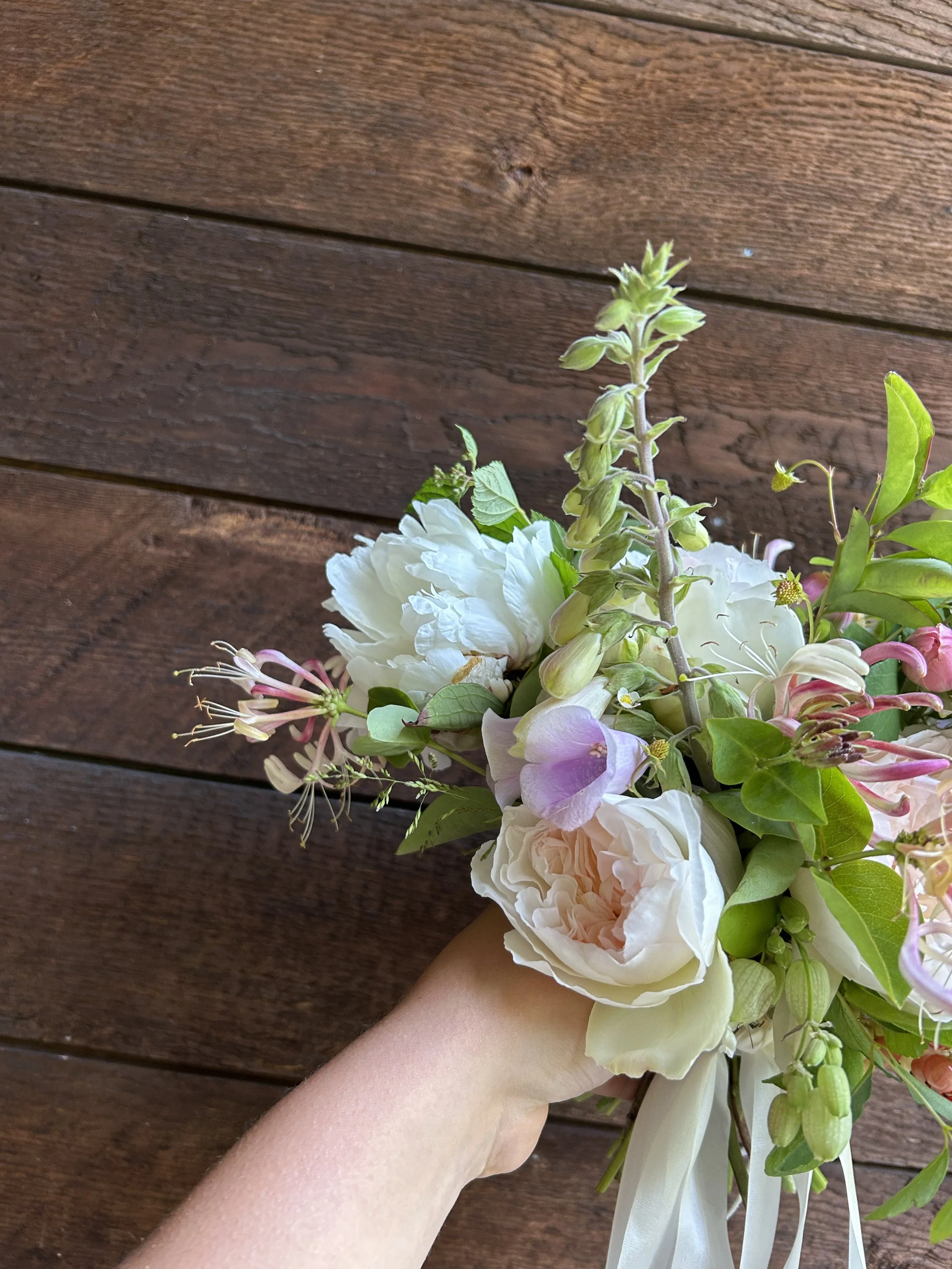 A hand holding a bouquet of pink and white flowers with green leaves against a wooden background.