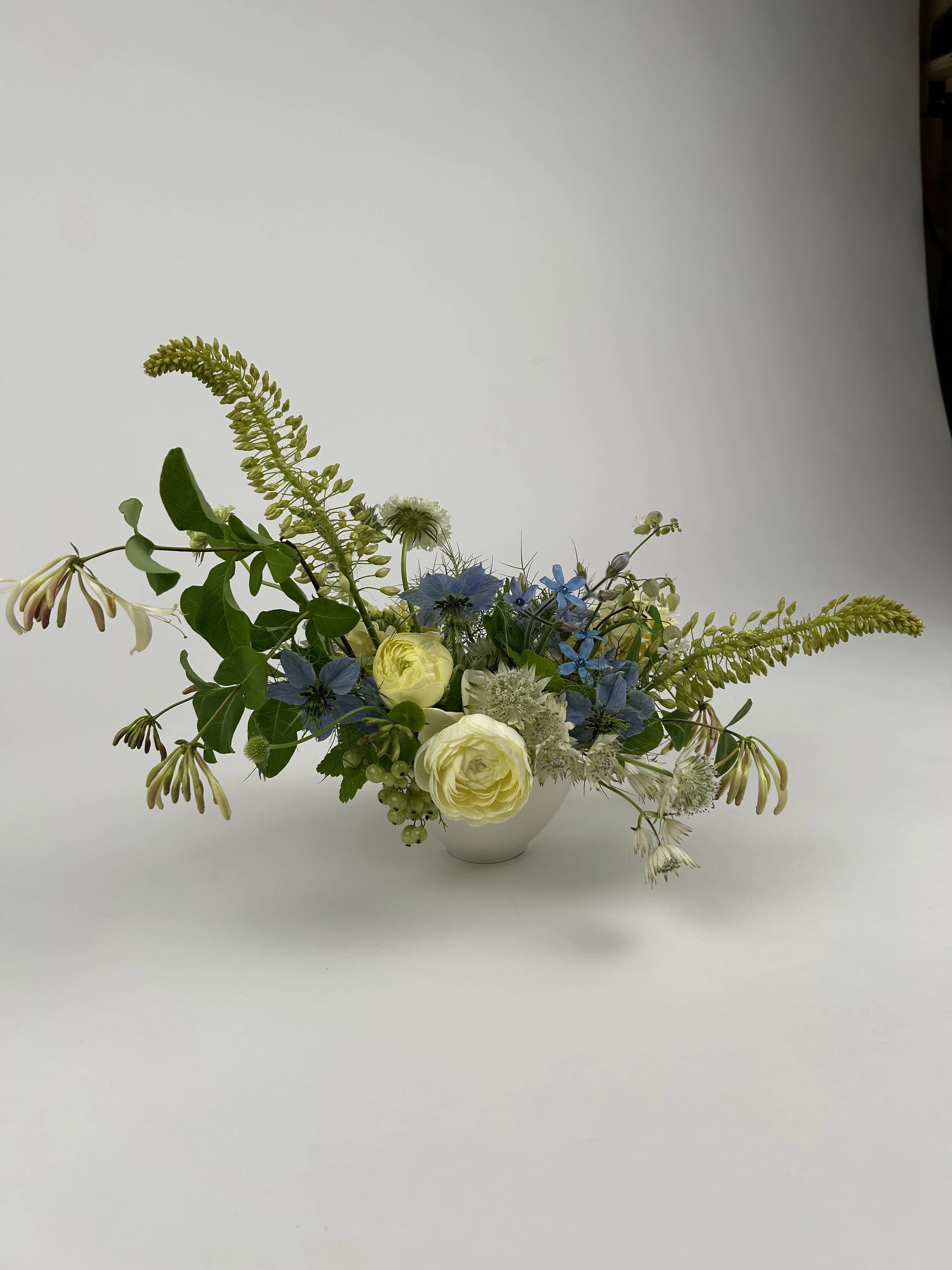 A floral arrangement in a white vase with white, yellow, blue, and green flowers and greenery on a plain white background.