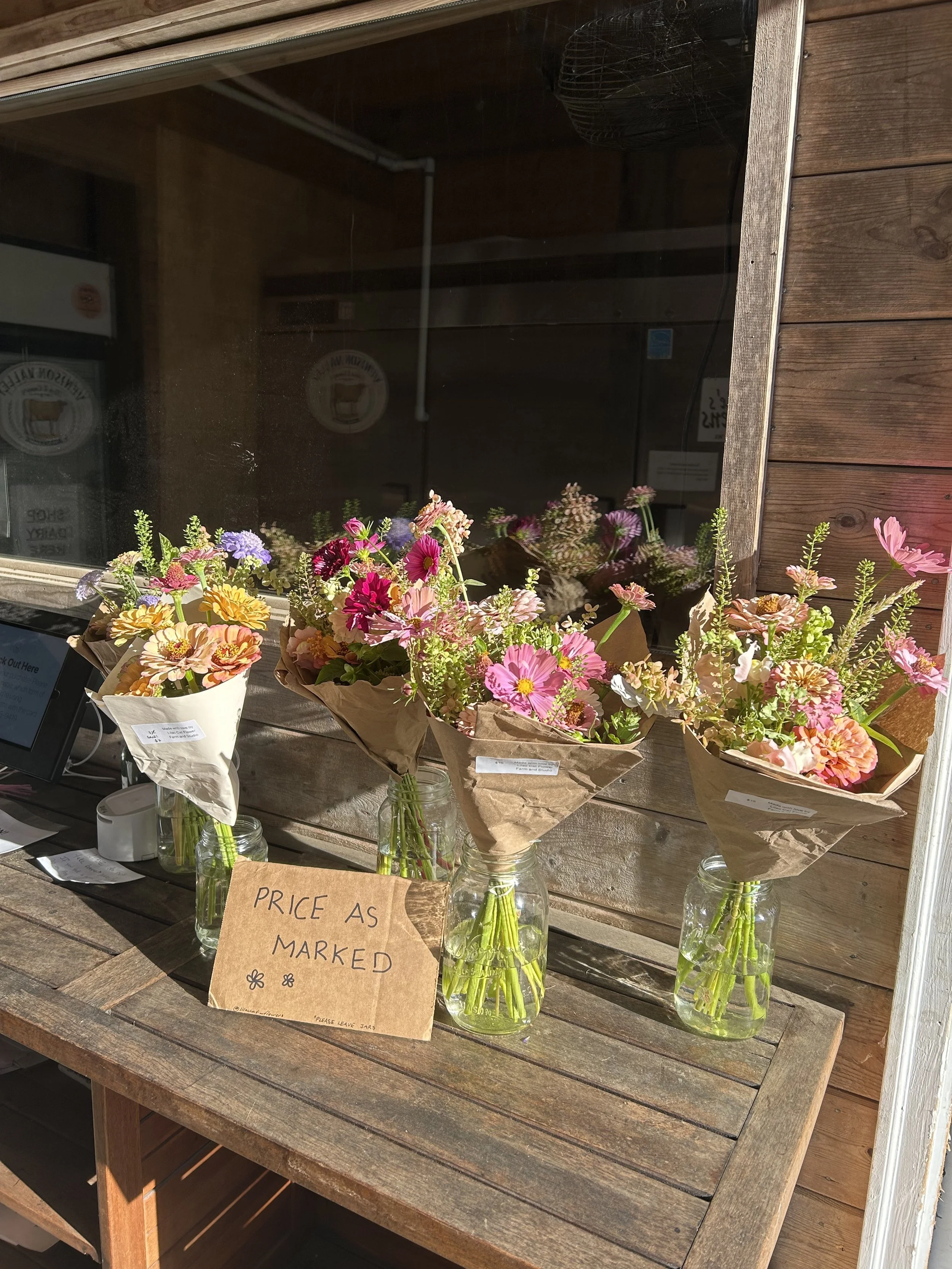 Three bouquets of colorful flowers in glass jars on a wooden table. A handwritten sign reads 'Price as marked.' The background shows a window reflecting more flowers and a wood-paneled wall.