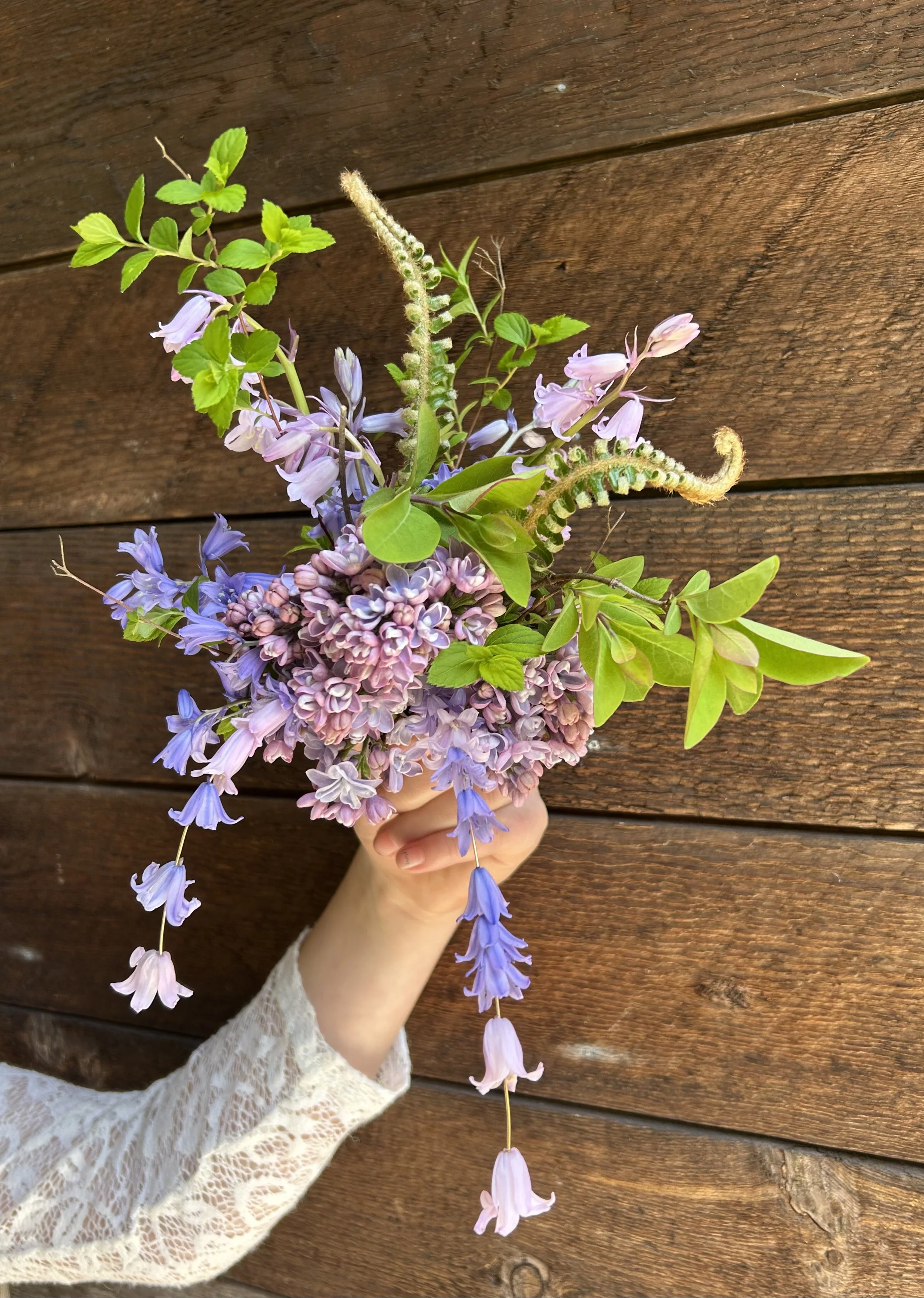 A hand holding a bouquet of purple and pink flowers with green leaves against a wooden background.