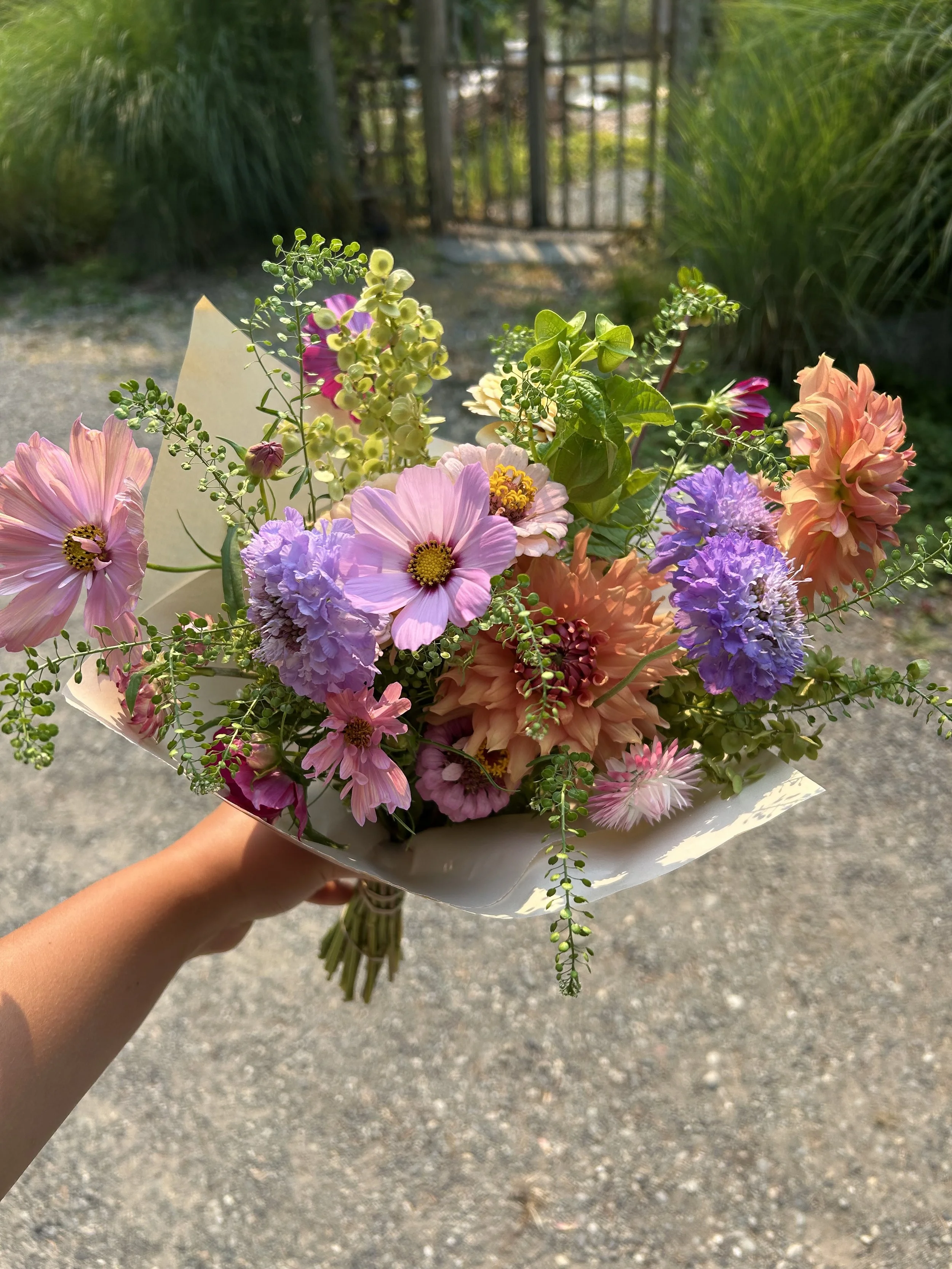 A person holding a colorful bouquet of various flowers including pink, purple, and peach blooms. The background shows a dirt pathway with a gate and greenery.