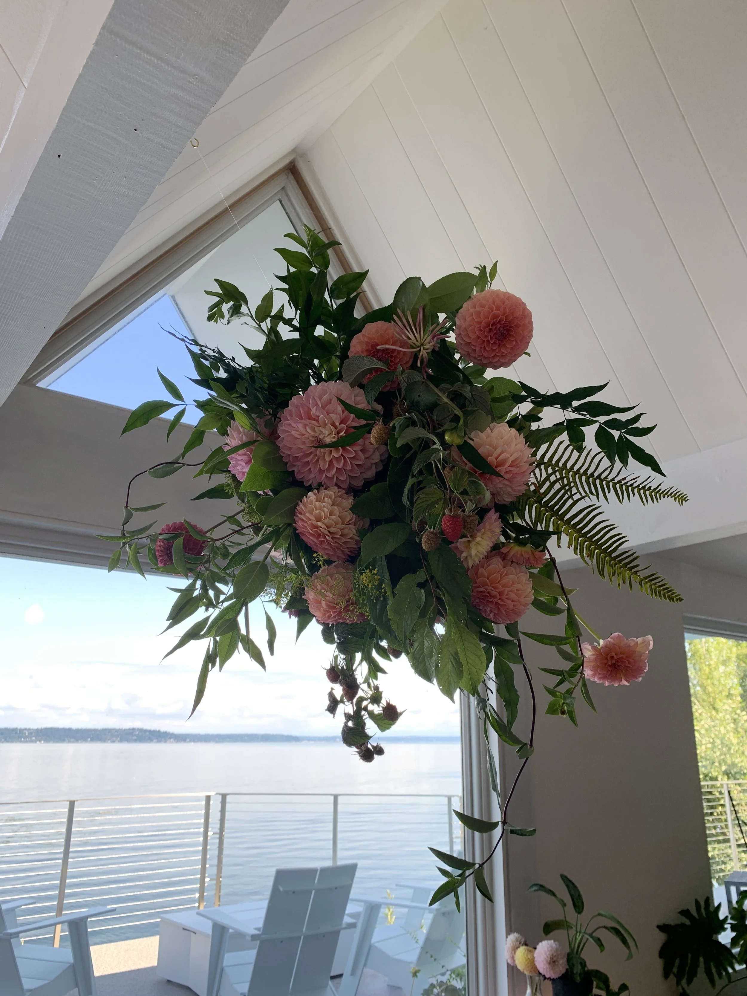 Hanging bouquet of pink dahlias, greenery, and ferns indoors near a large window overlooking a body of water.