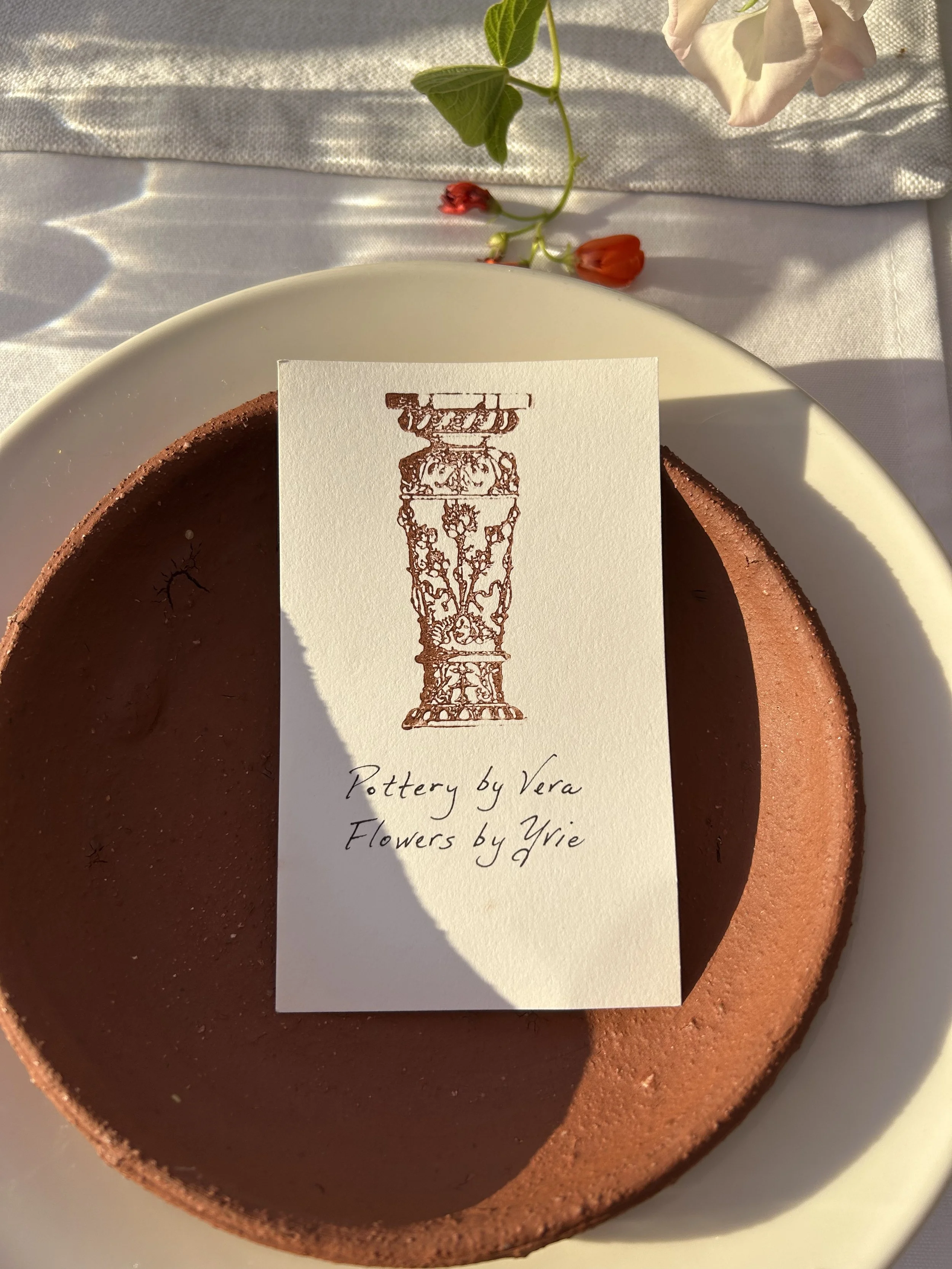A round, reddish-brown ceramic pot with a smooth interior, placed on a white plate with a business card on top. The card has a drawing of an intricate tall, decorative vase and the text 'Pottery by Vera, Flowers by Yrie'. There are some green leaves 