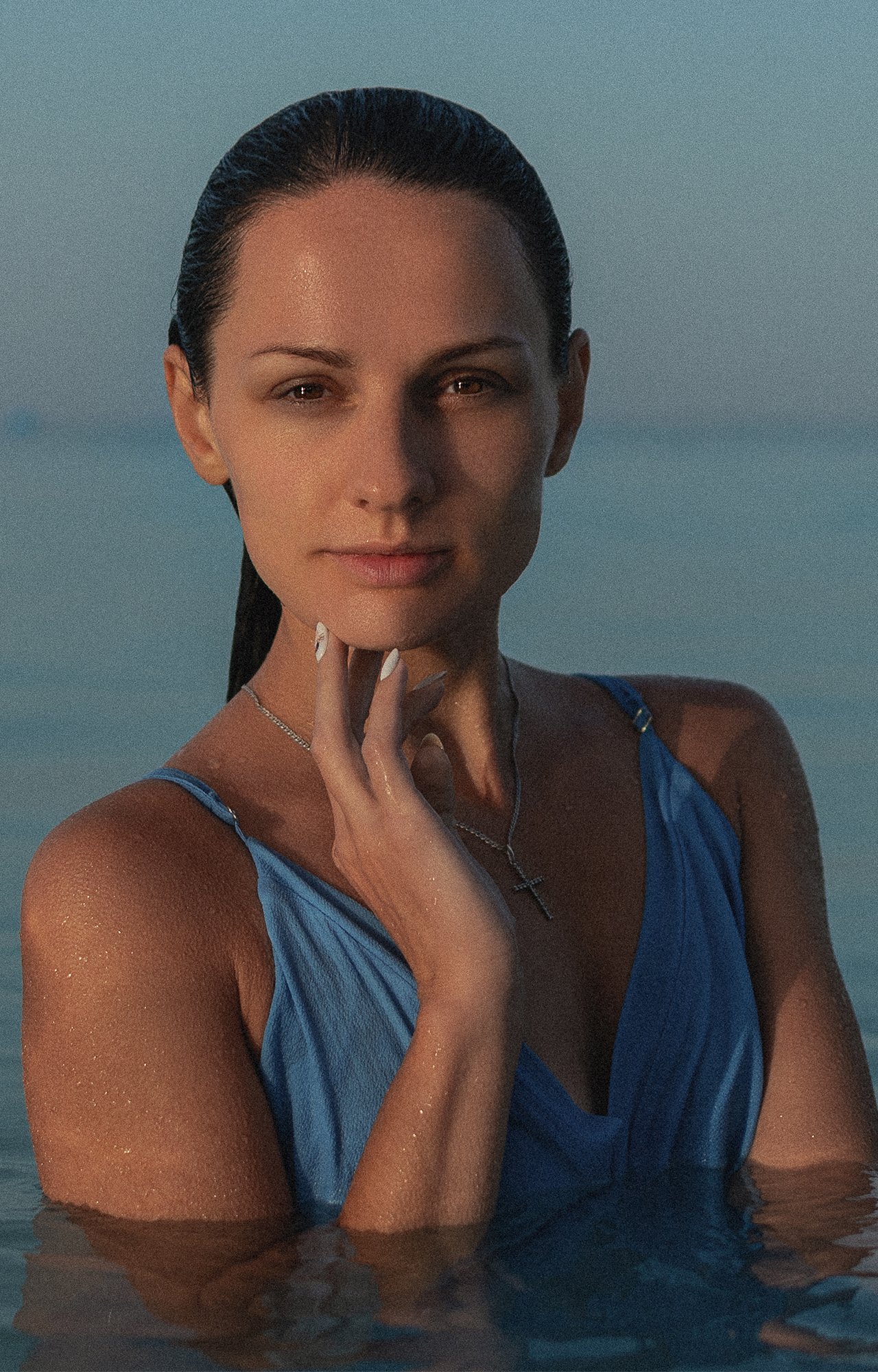 Mujer joven en el agua con cabello recogido, ropa azul, posando con expresión confiada, en un entorno natural acuático durante el atardecer o amanecer.