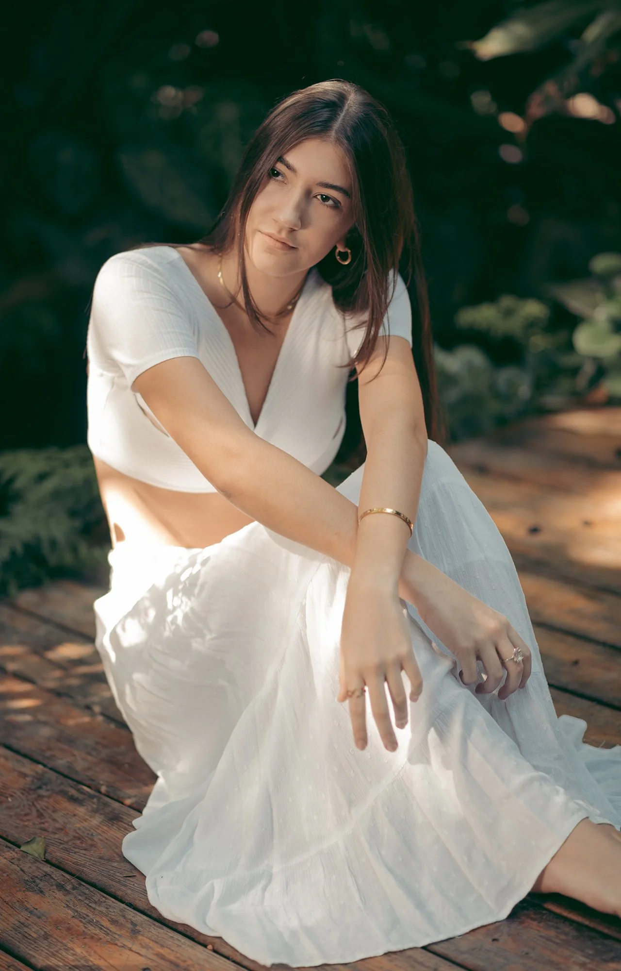 Mujer con cabello largo y oscuro, vistiendo blusa blanca y falda larga, sentada en un muelle de madera en un entorno natural con sombra de árboles.