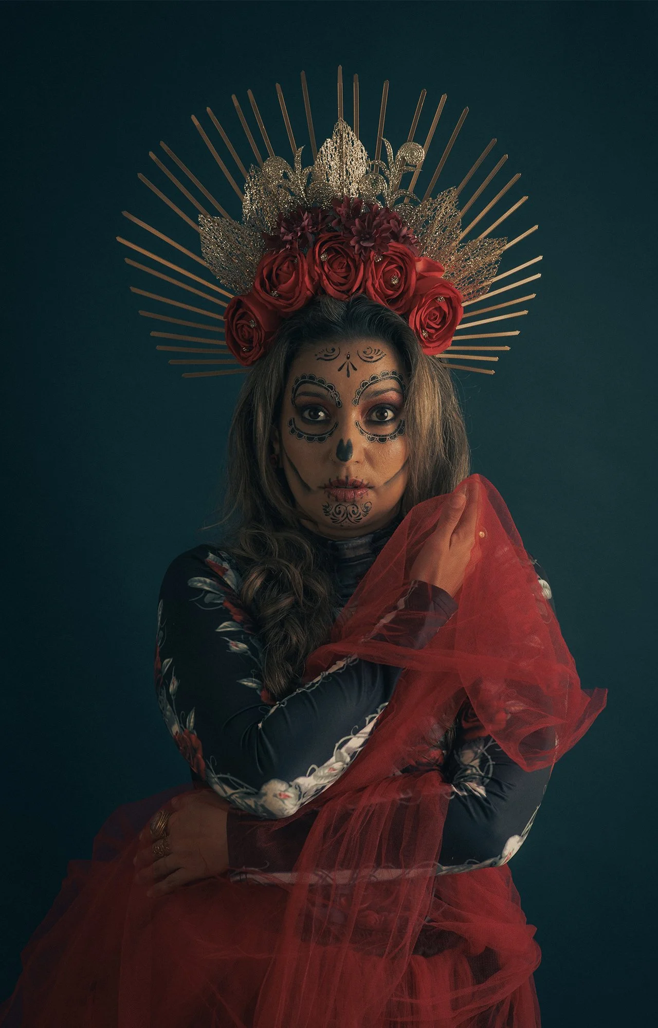 Mujer con maquillaje de calavera, coronada con rosas rojas y detalles dorados, sosteniendo tela roja, contra fondo oscuro.