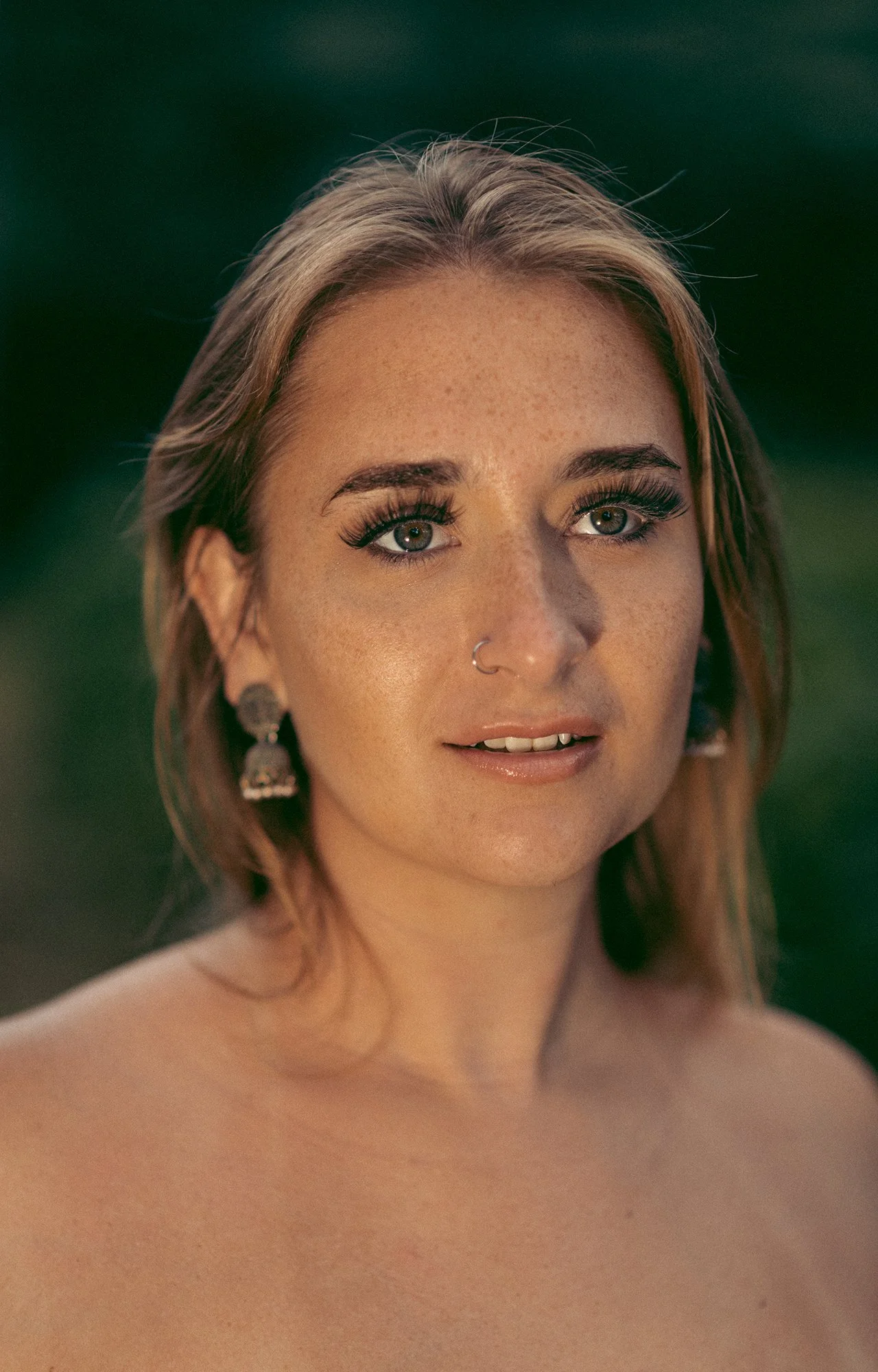 Retrato de una mujer con cabello rubio, pecas y ojos claros, usando aretes y un piercing en la nariz, con fondo difuso y natural.