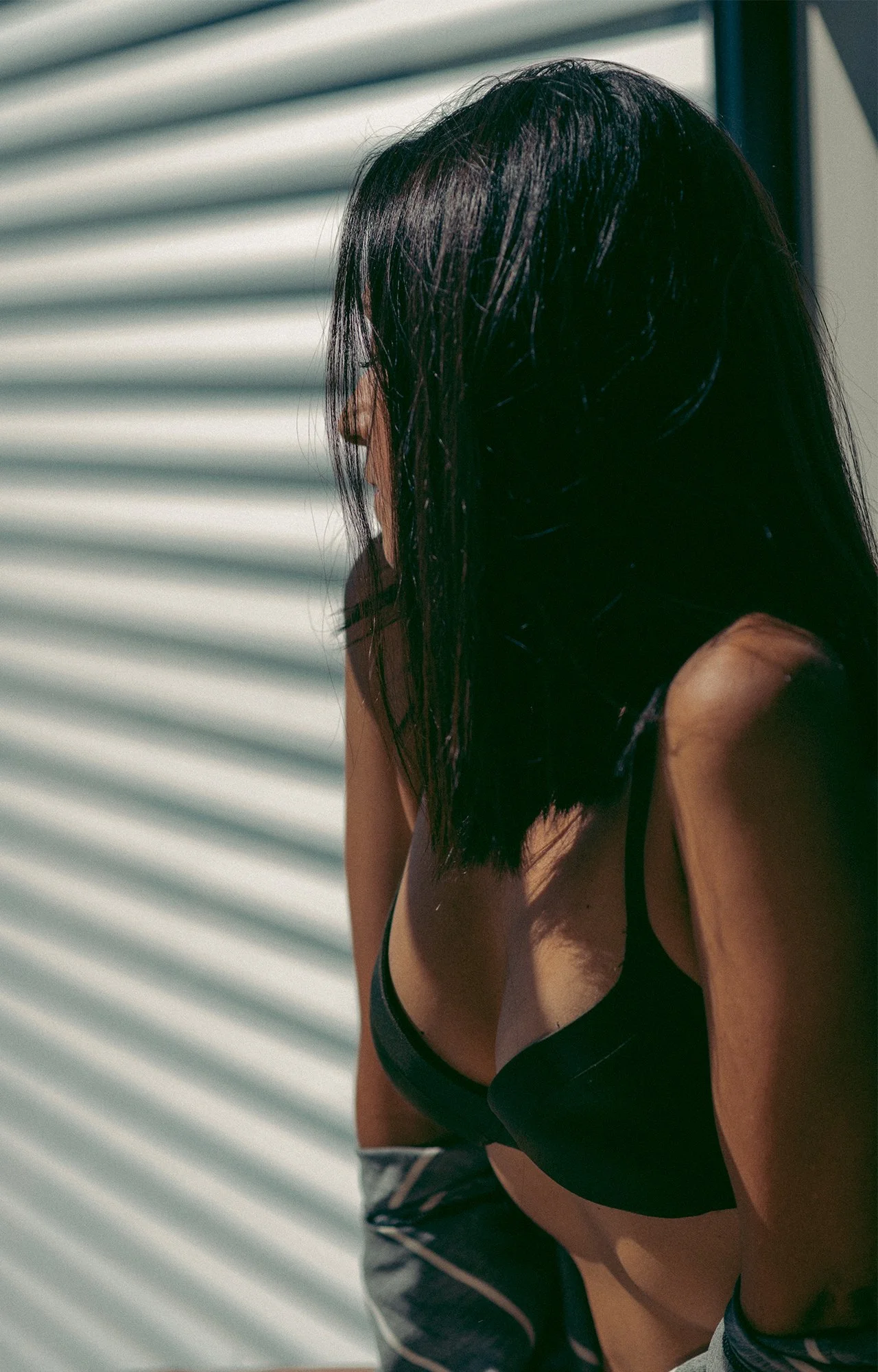 Mujer de cabello negro y piel bronceada, con top negro, de espaldas, apoyada contra una pared con líneas horizontales en un entorno soleado.