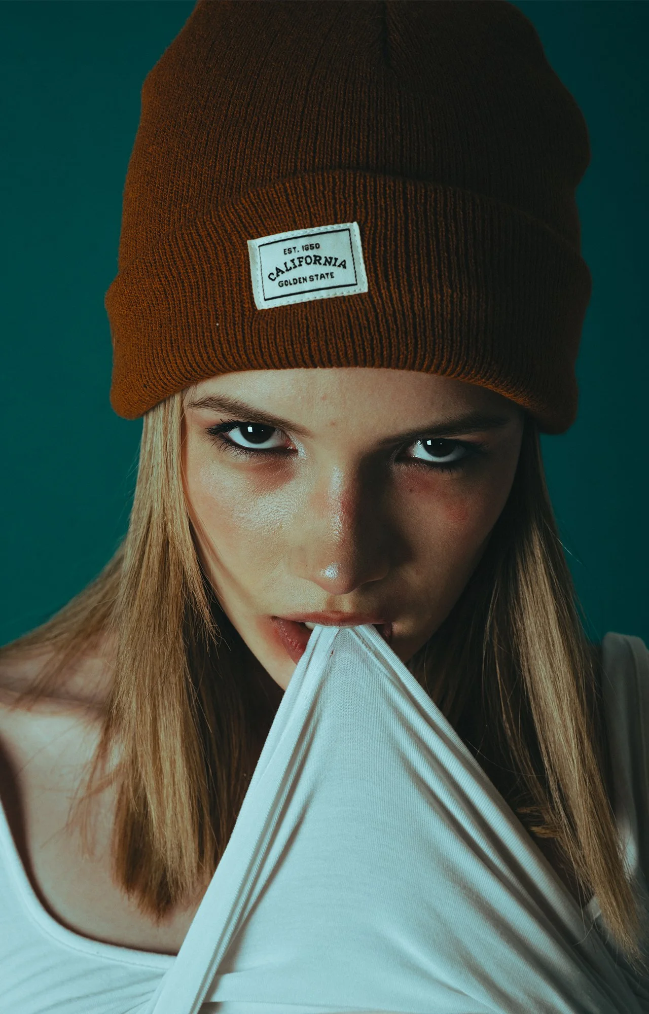 Joven mujer con gorro marrón y camiseta blanca, mirando intensamente a la cámara, tocando la camiseta con la boca.