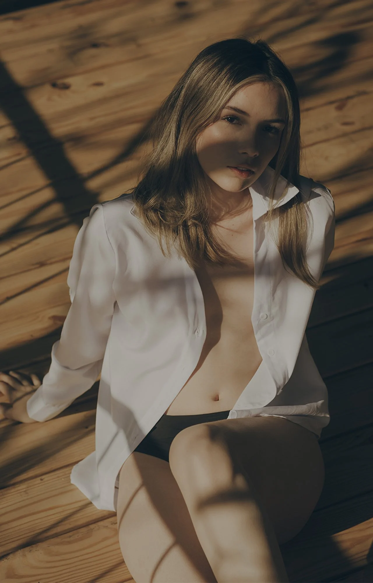 Juvenil mujer con cabello rubio, semidesnuda, con camisa blanca abierta, sentada en una superficie de madera, con sombras creadas por la luz y objetos que no se distinguen claramente.