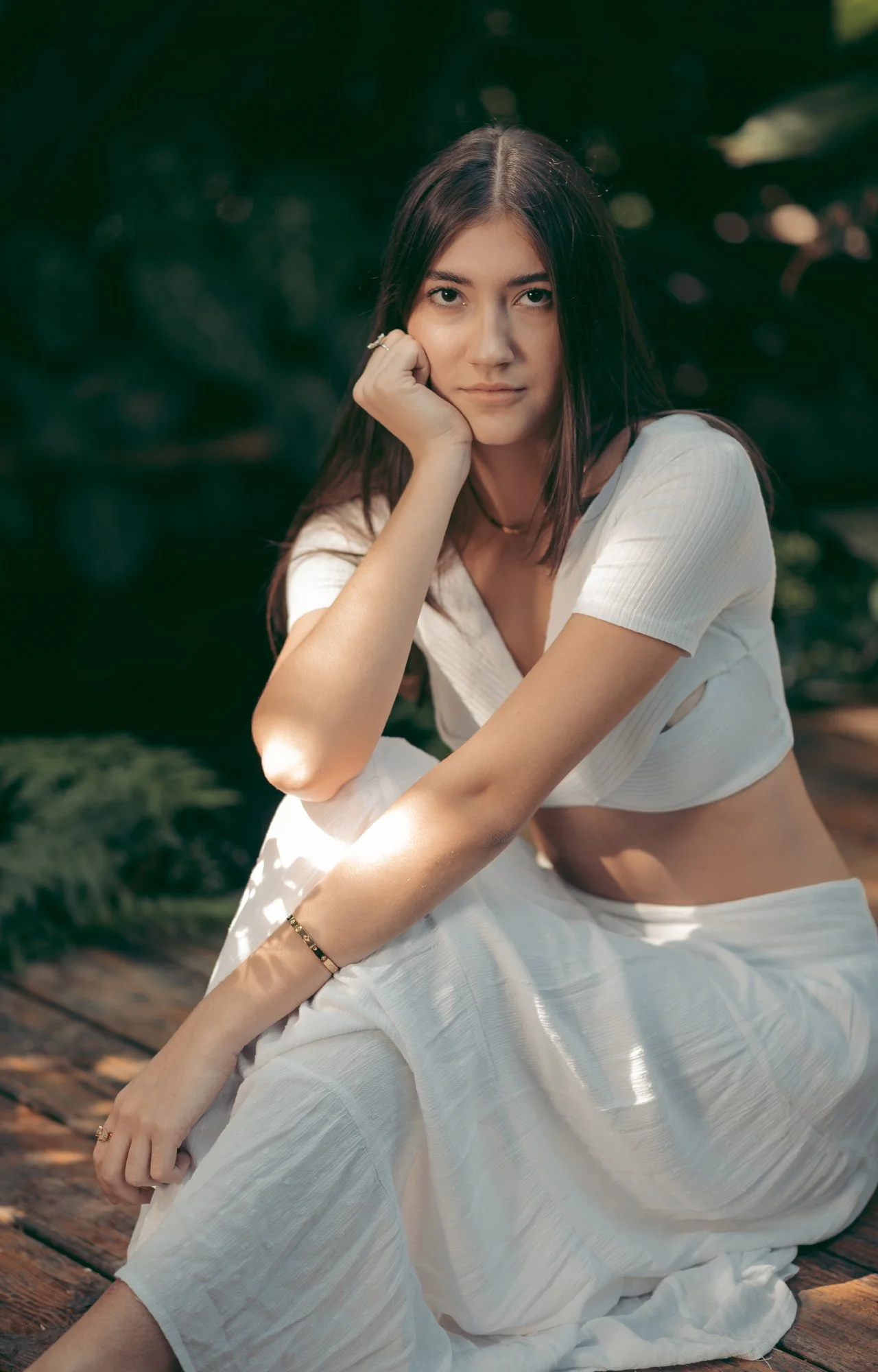 Joven mujer sentada en un muelle de madera en un entorno natural, con cabello oscuro, ropa blanca y expresión pensativa.