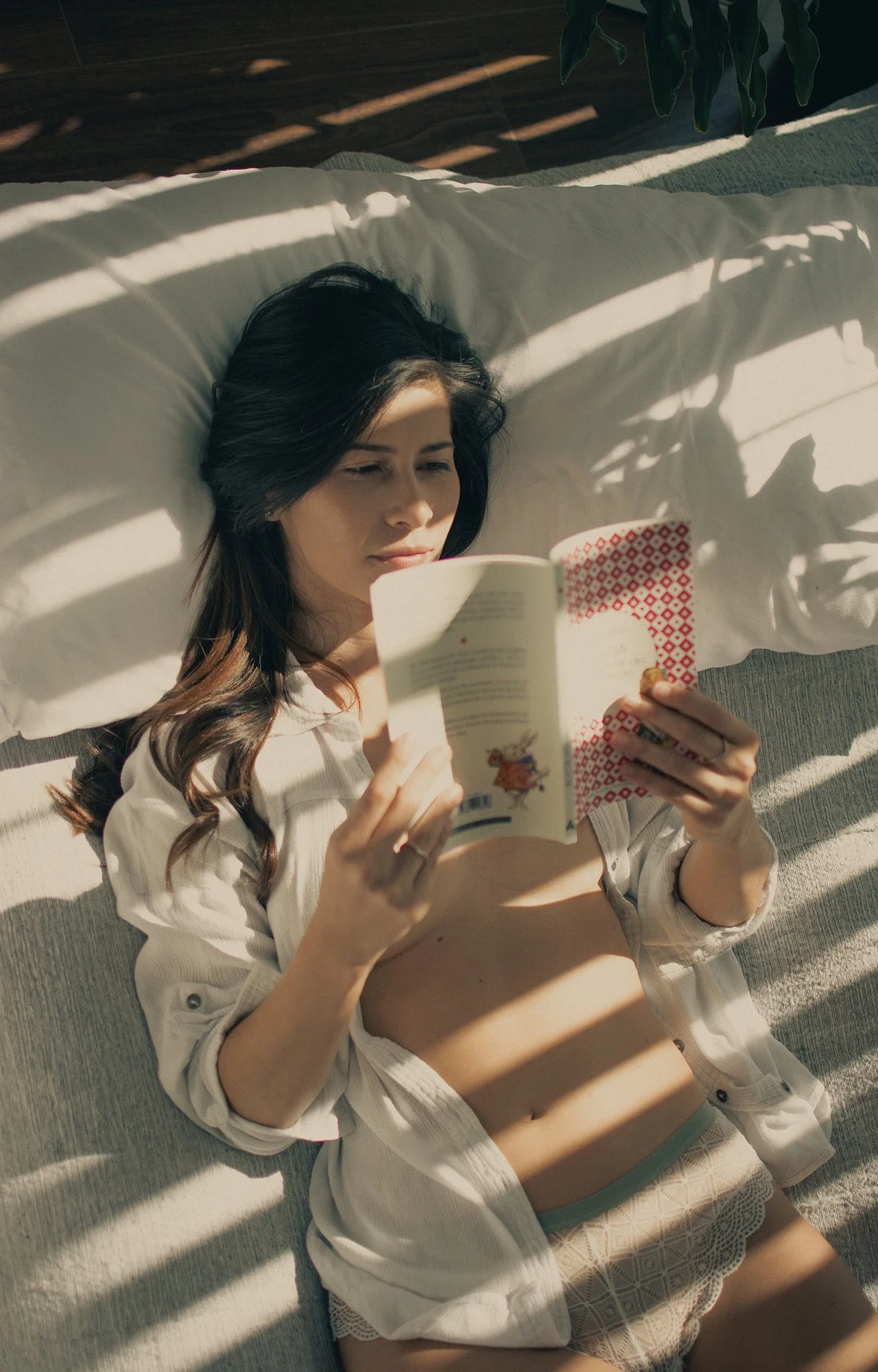 Joven leyendo un libro mientras está acostada en un sofá, con sombras de plantas que entran por una ventana, en un ambiente soleado y tranquilo.