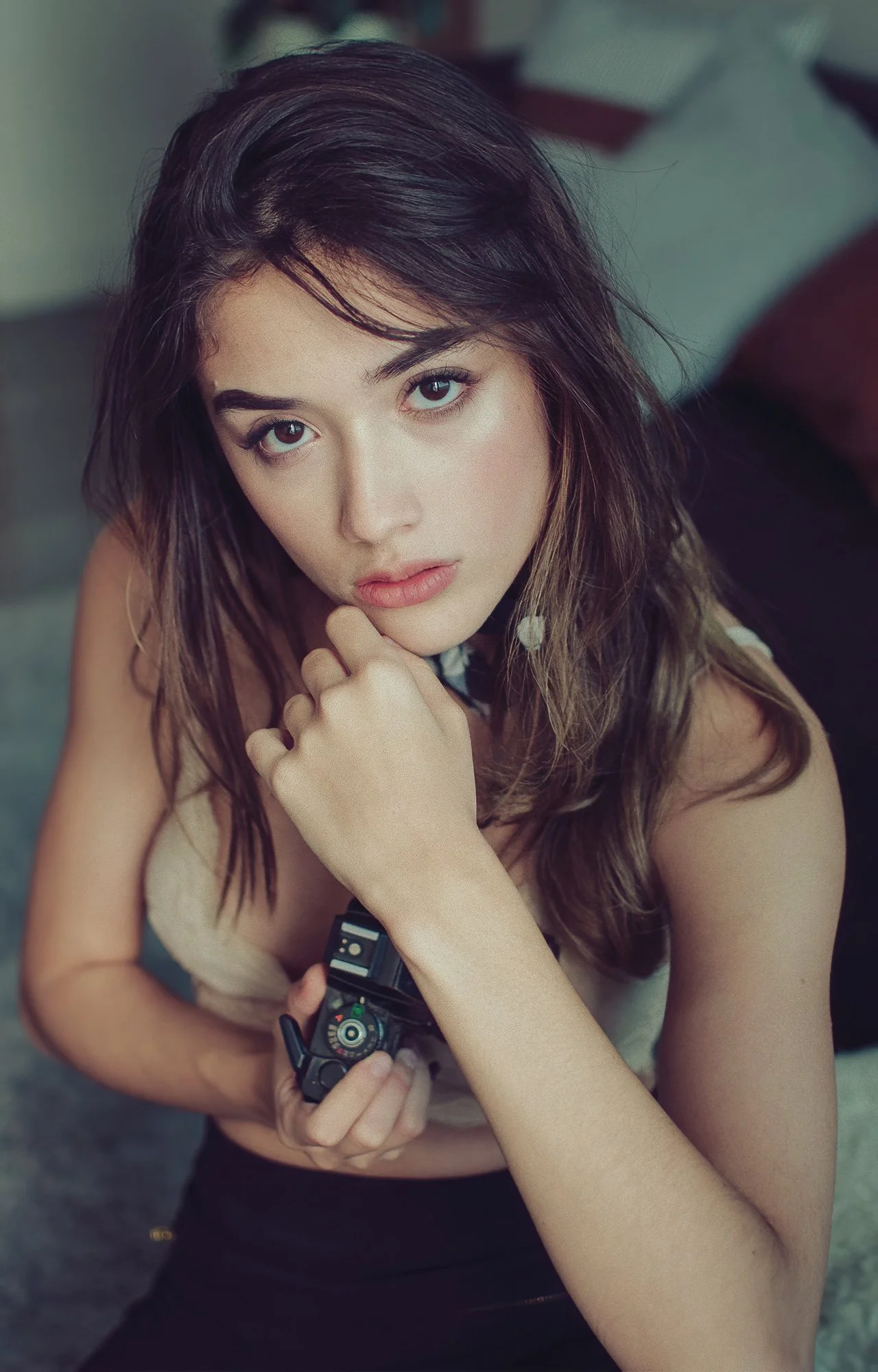 Una joven con cabello castaño oscuro y ojos color azul mira hacia la cámara, sosteniendo una cámara en su mano izquierda, con una expresión pensativa.