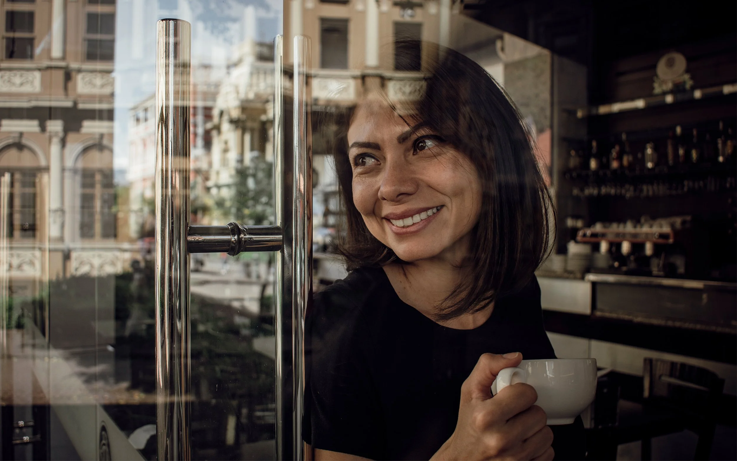 Mujer sonriendo con una taza de café, vista a través de una ventana de un café con edificios en la calle