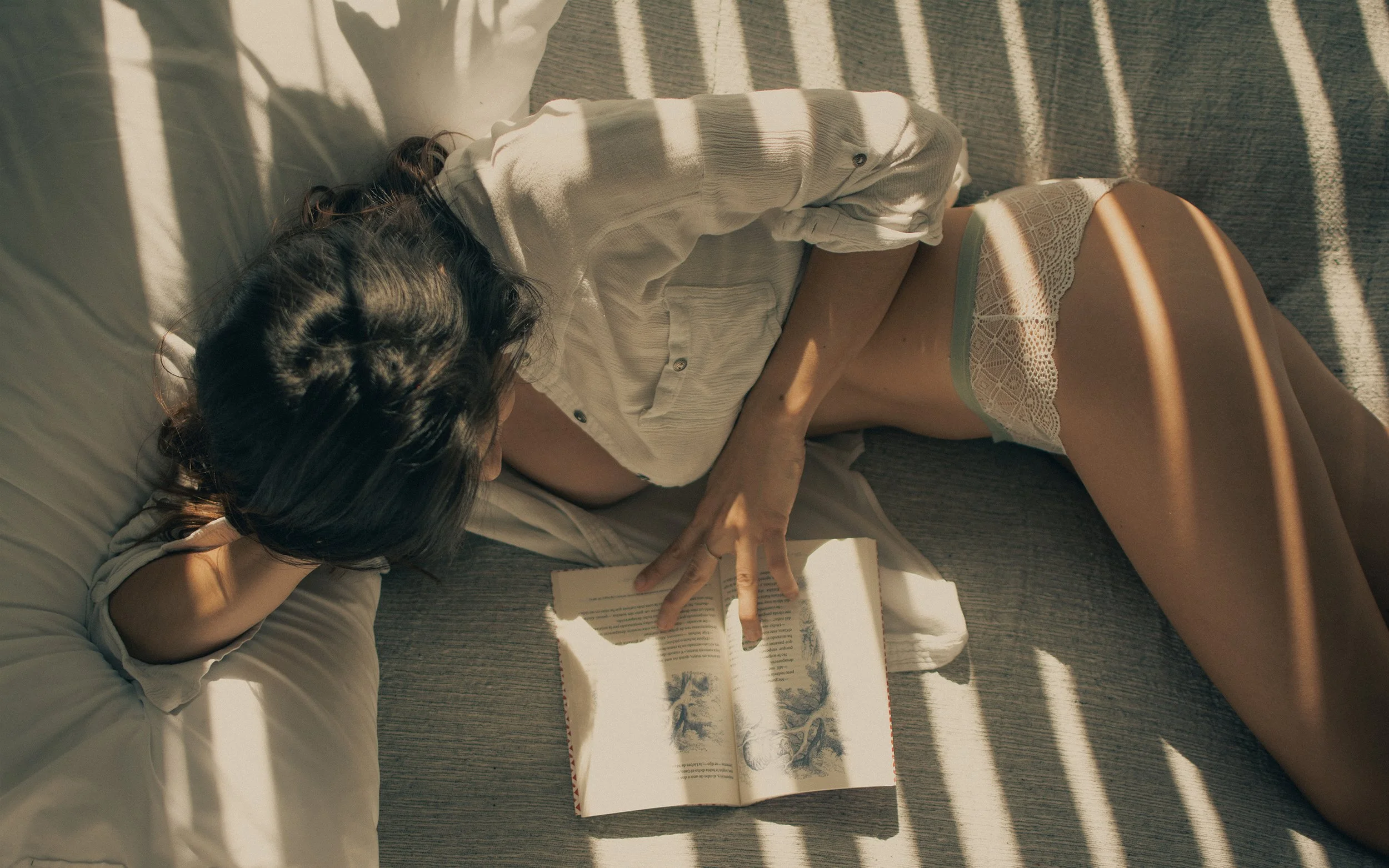 Mujer recostada en la cama, leyendo un libro, con sombra de persianas.
