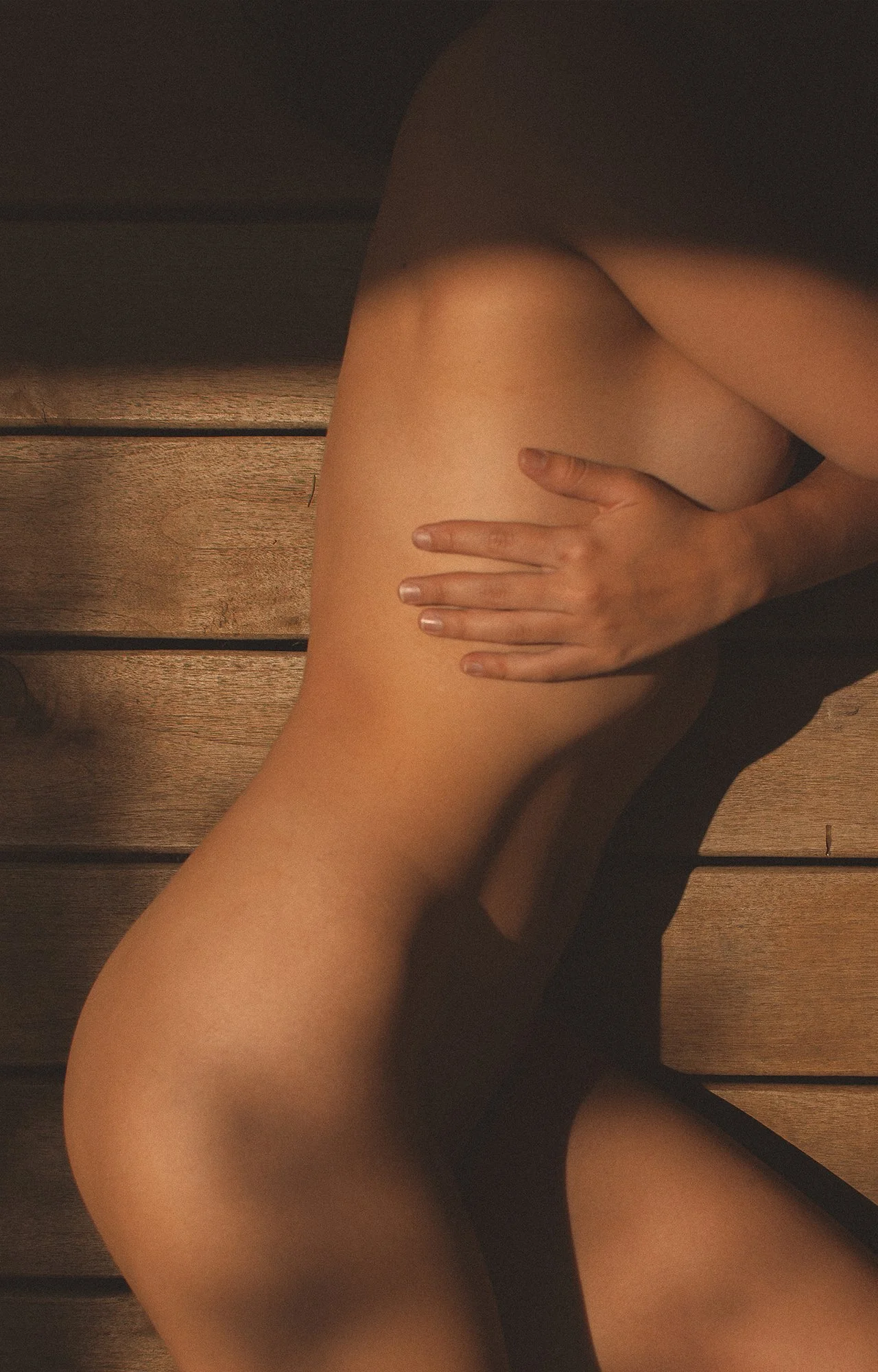 Una persona desnuda recostada sobre una superficie de madera, cubriéndose parcialmente con una mano.