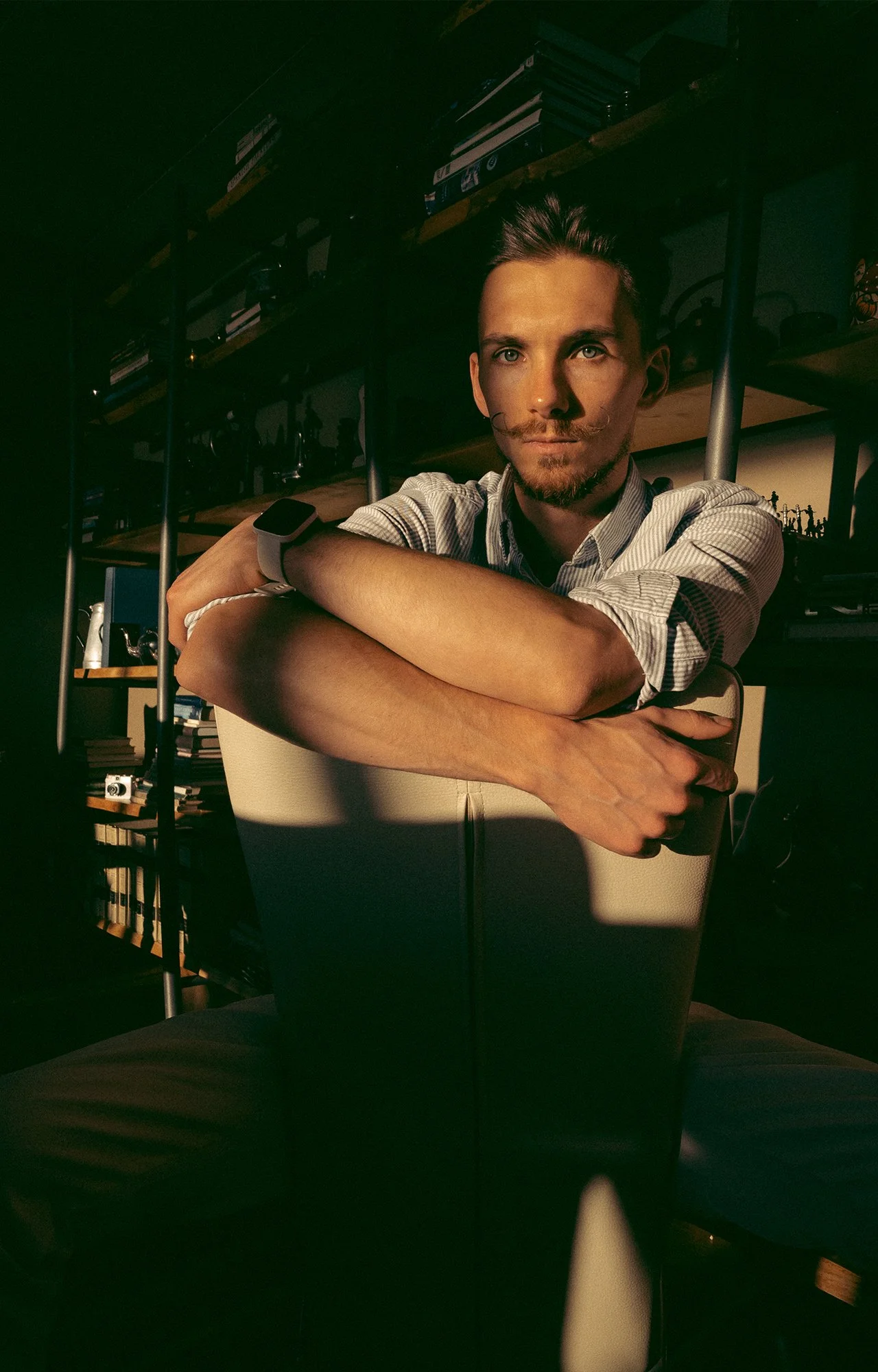 Un hombre con barba y bigote, con cabello recogido, sentado en una silla, en un ambiente con estanterías de libros y objetos en el fondo, iluminado por luz cálida.