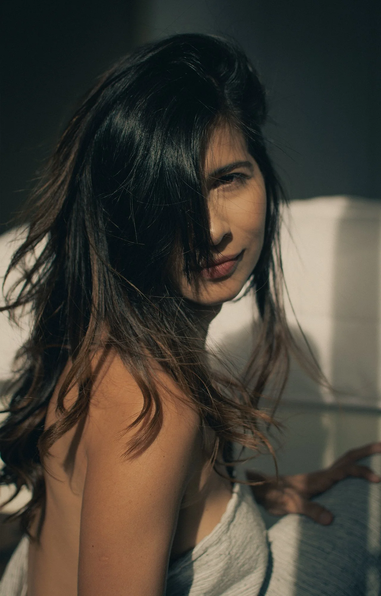 Mujer con cabello largo y oscuro parcialmente cubriendo su rostro, sonriendo ligeramente, en un ambiente con luz natural y fondo oscuro.