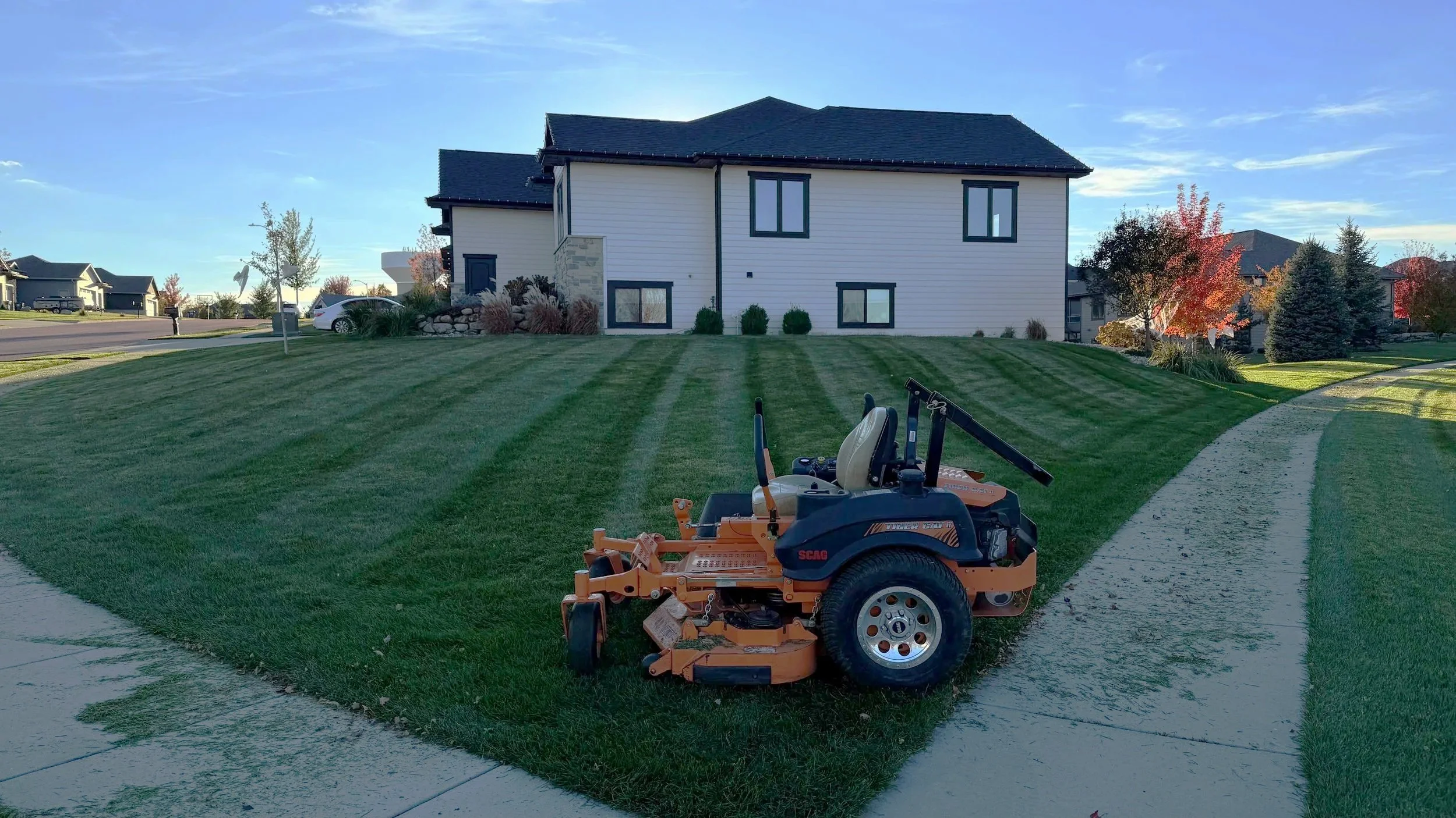 A lawn mower parked on freshly mowed grass in front of a white house with dark roof in a suburban neighborhood with trees and other houses in the background.