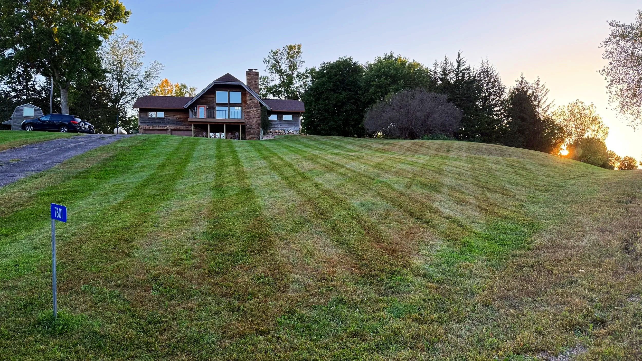 A house with large glass windows at the top of a sloped, freshly mowed lawn during sunset, with trees in the background and a blue street sign reading '760' in the foreground.