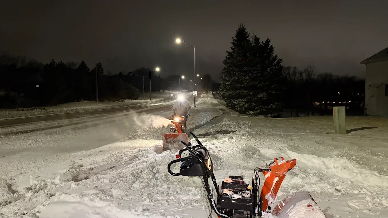 Two crew members clearing snow with snow blowers at a commercial property before dawn