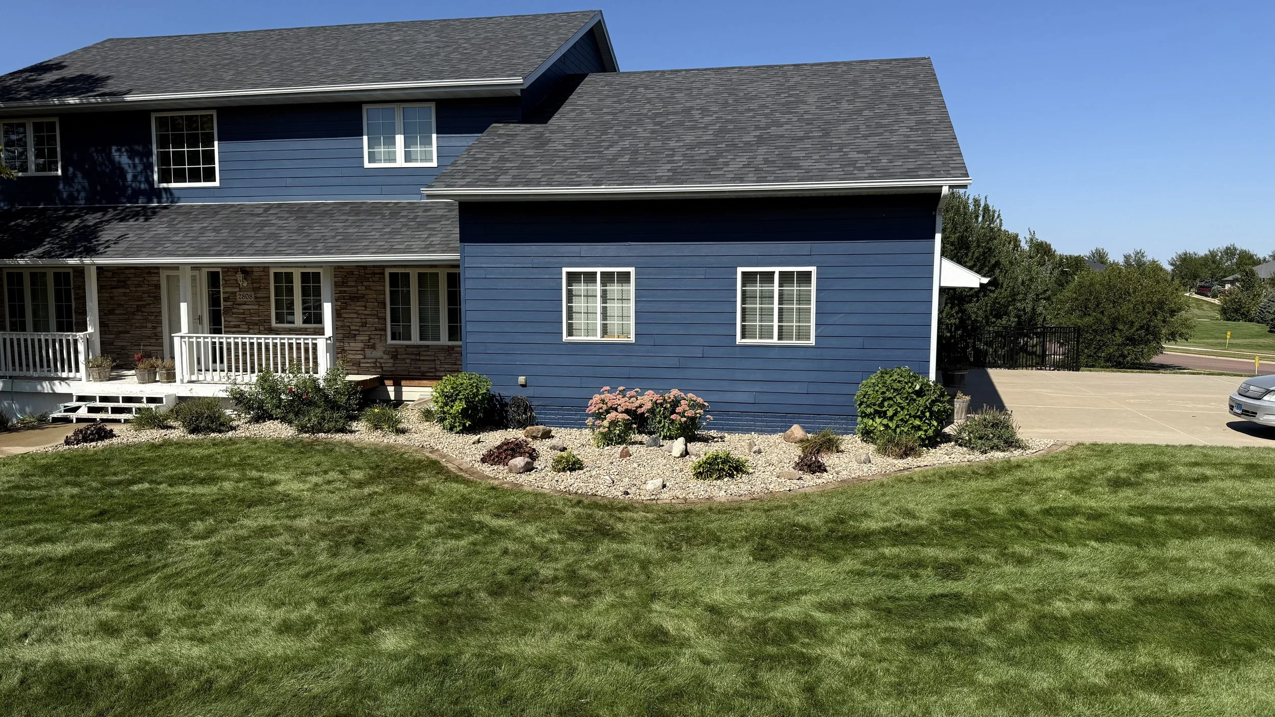A house with blue siding and a gray shingle roof, featuring three windows on the blue section. The front yard has a well-maintained green lawn and a landscaped garden bed with flowers, rocks, and bushes in front of the house. There is a white porch w