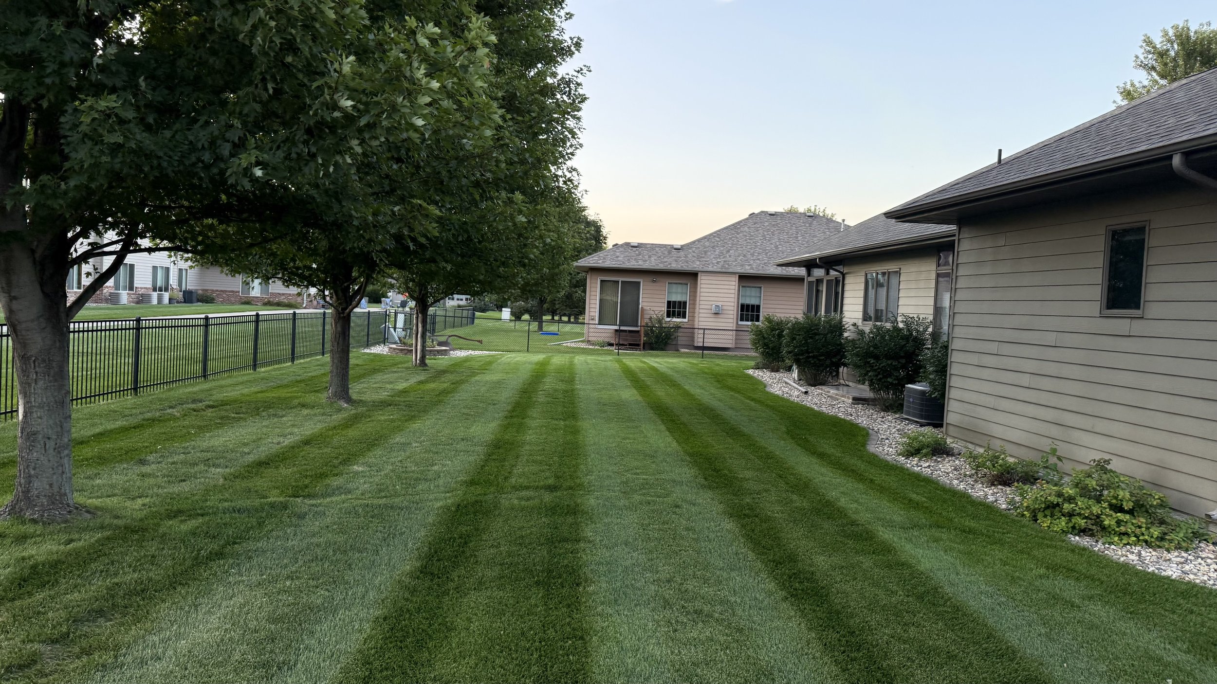 Well-maintained backyard with striped green lawn, trees, and houses with outdoor units and gardens.
