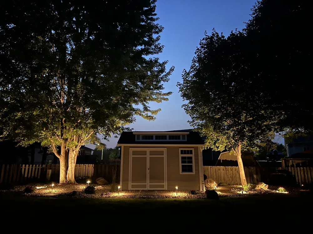The same backyard shed and rock bed at dusk, lit by low-voltage path lights and tree uplighting