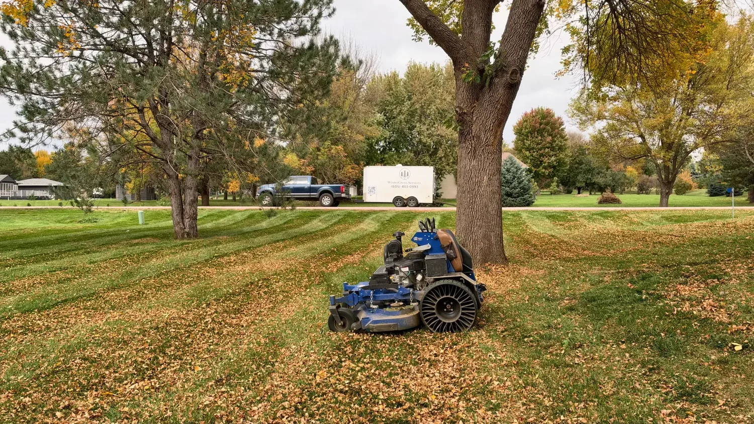 Stand-on mower beside a tree on a leaf-covered lawn, with the WinterGreen Services truck and trailer parked behind