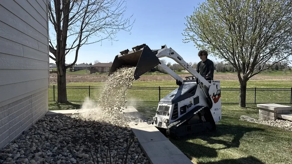 Crew member operating a compact track loader to pour landscape rock into a bed beside a home