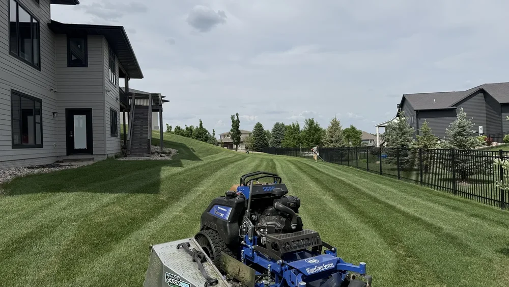 Mower with clipping catcher on a striped backyard lawn as a crew member trims along the fence