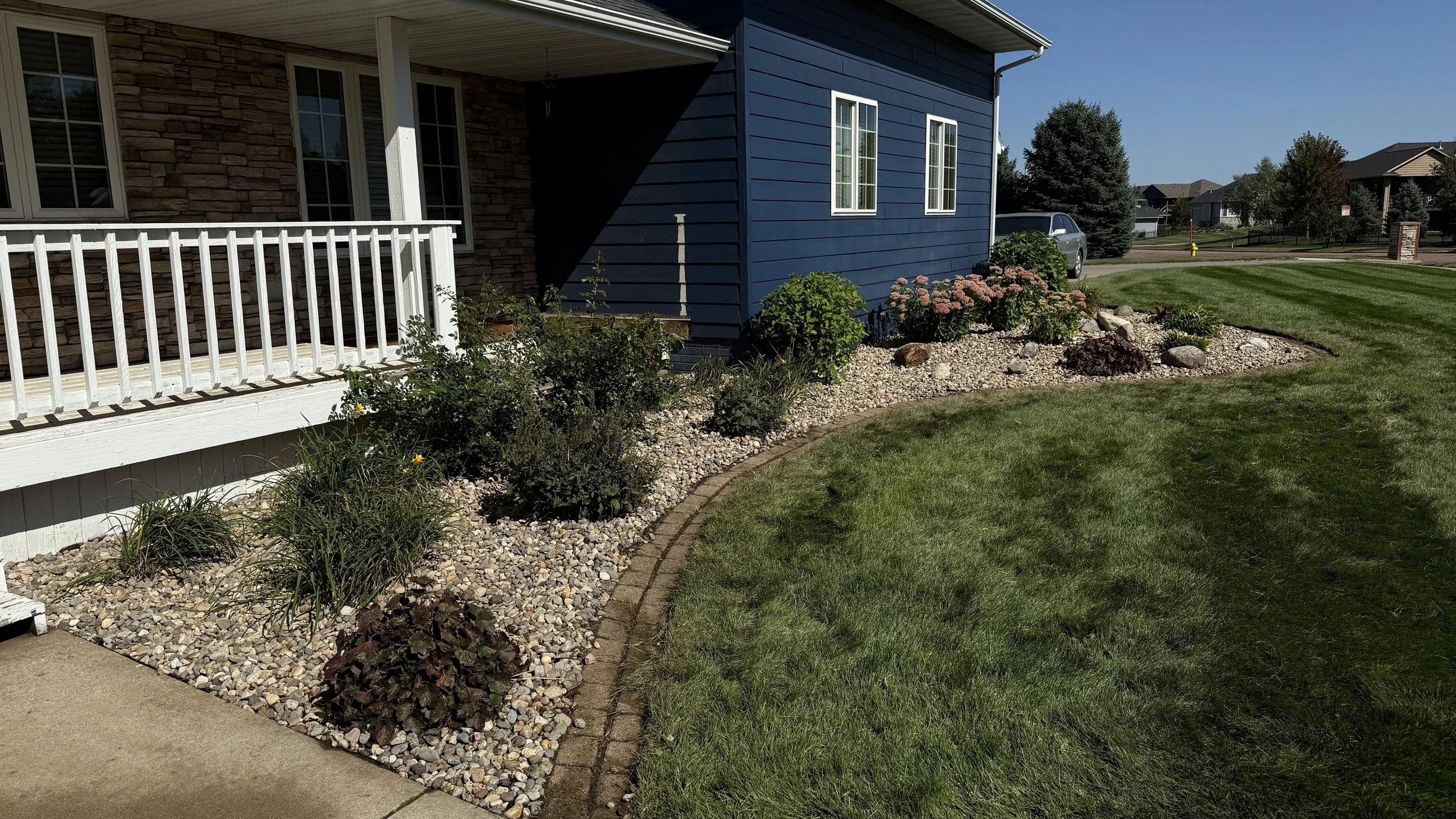 View of a residential yard with a garden bed edged with bricks, filled with various plants and flowers, next to a house with a stone and blue siding exterior, white railing, and a porch. A grassy lawn extends into the distance with other houses and t