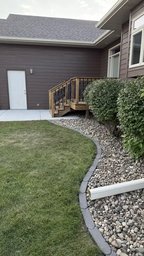 Curved paver edging separating a green lawn from a rock bed beside a wooden deck stairway