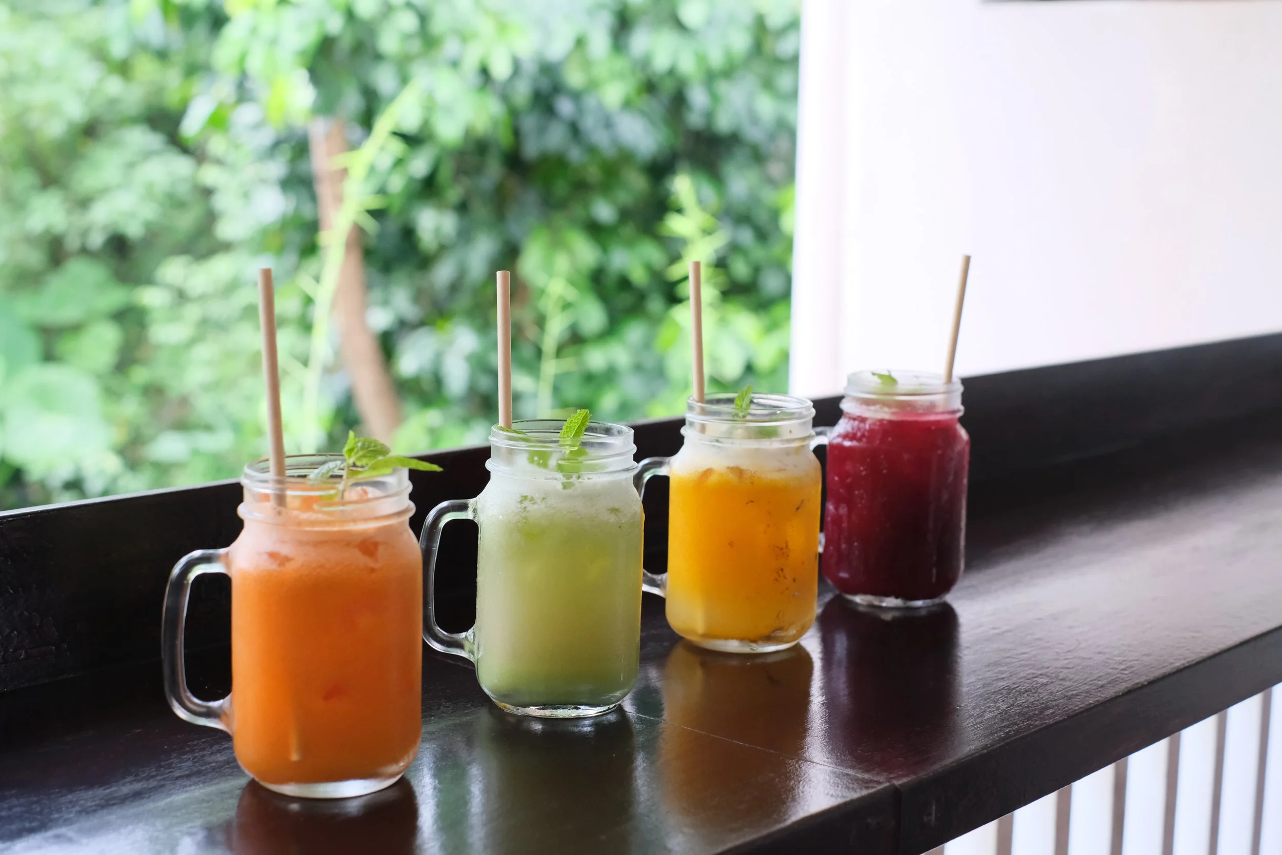 Four mason jar glasses with colorful drinks, garnished with mint leaves and straws, on a dark wooden surface with green foliage in the background.