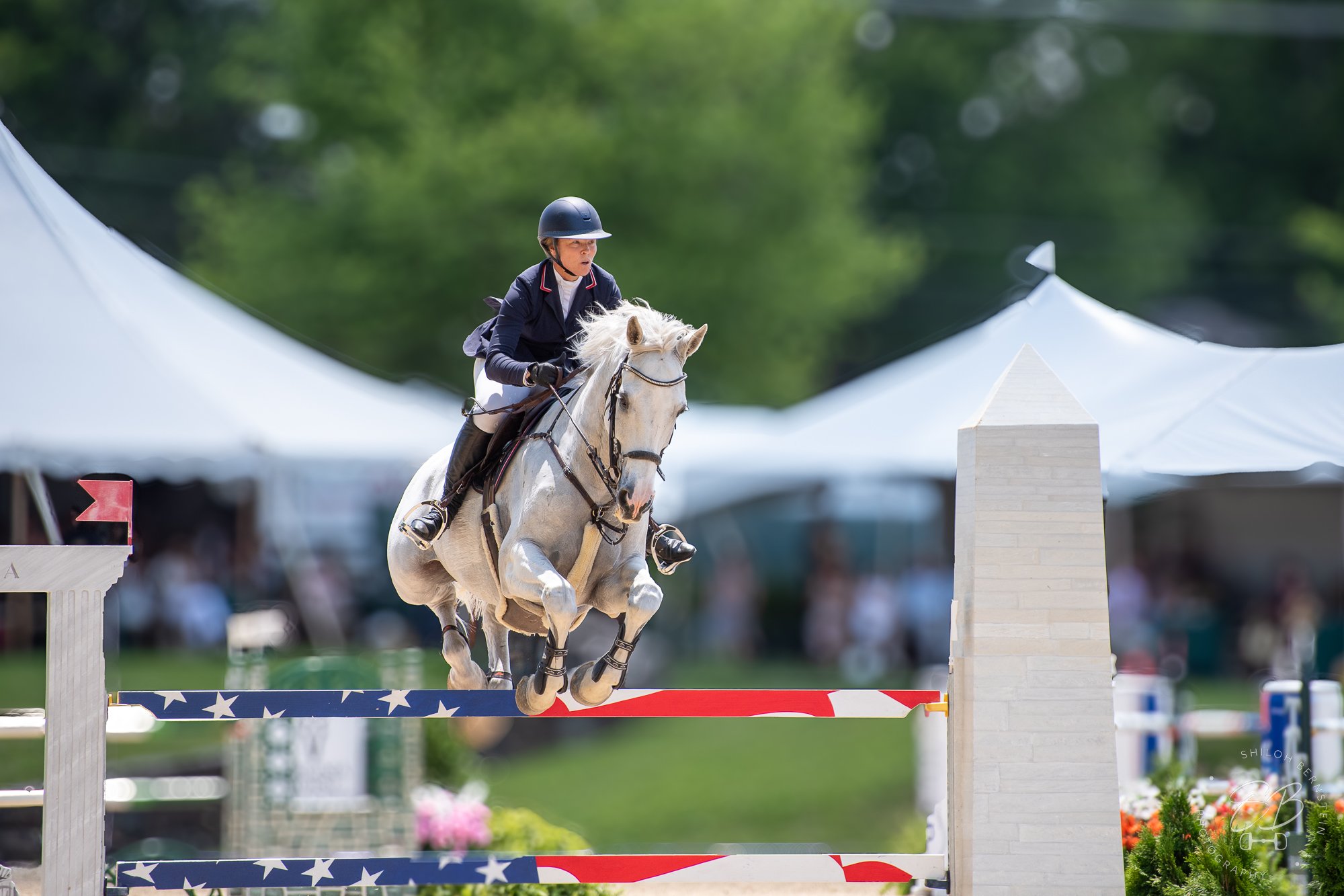 A young female equestrian in riding gear and a helmet riding a white horse over an obstacle with an American flag design during a show jumping competition, with tents and green trees in the background.