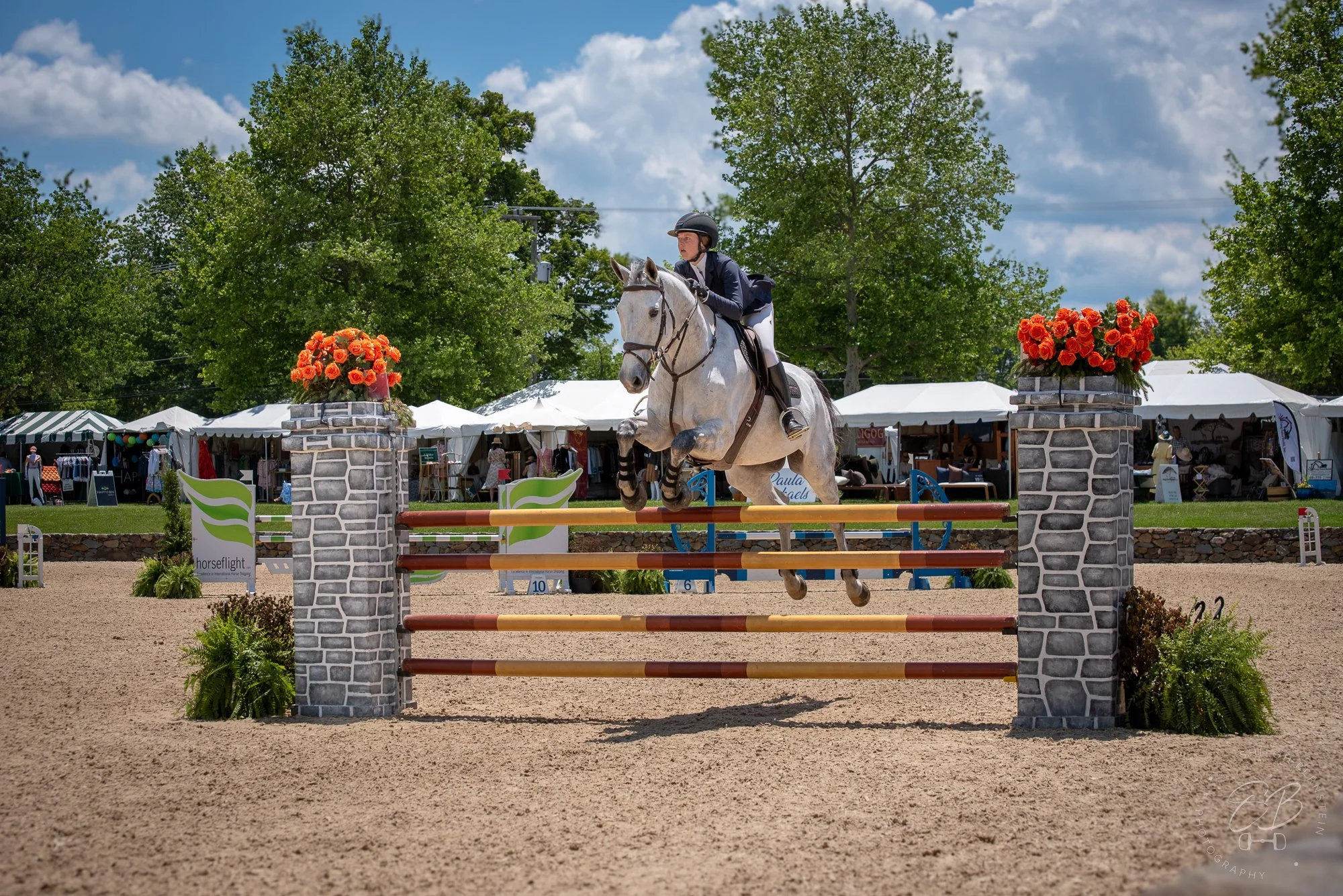 A female equestrian riding a gray horse over a show jumping obstacle at an outdoor competition, with tents, trees, and blue sky in the background.